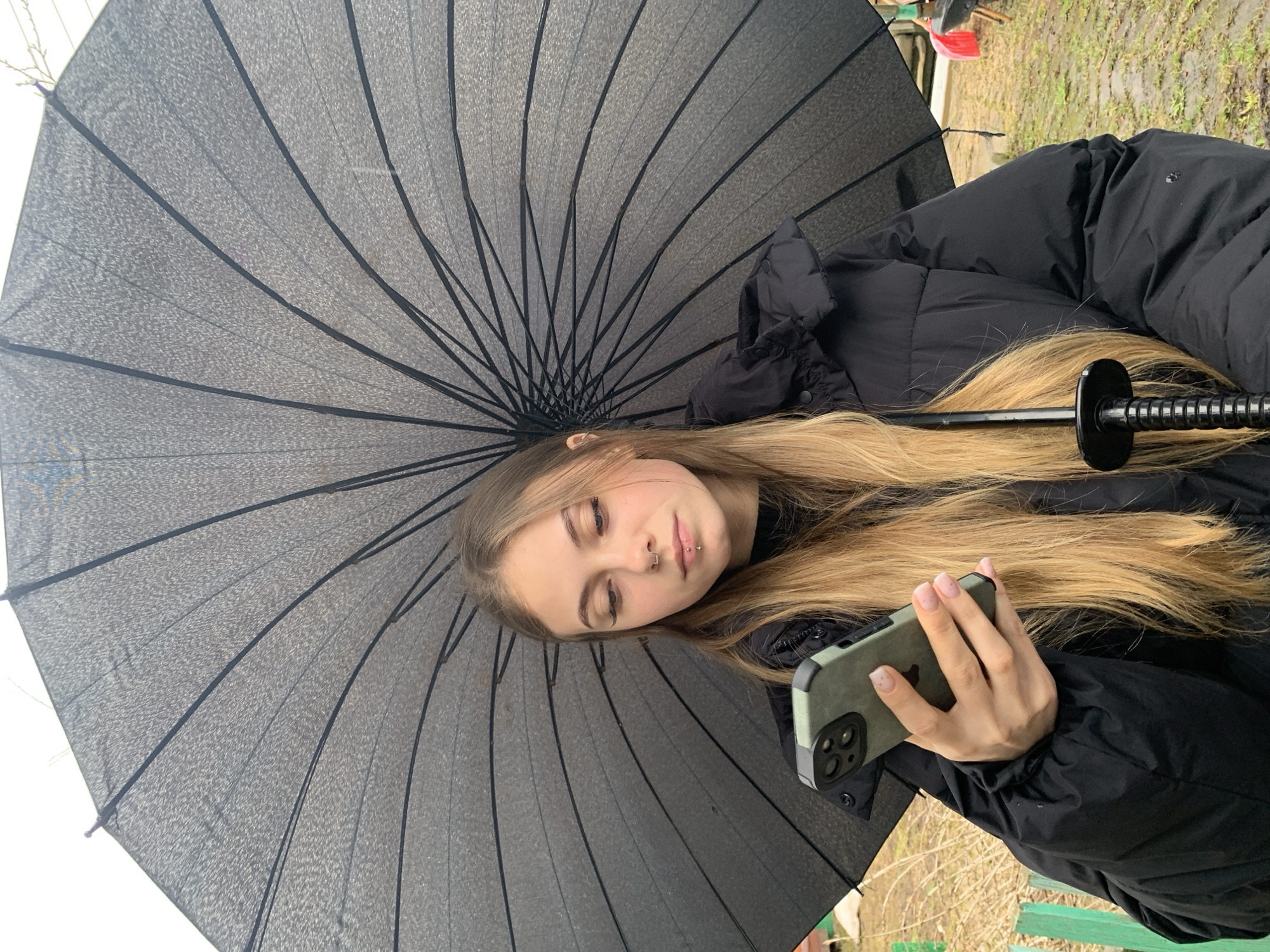 A young woman with long blonde hair is holding a smartphone in her right hand under a large black umbrella. The woman has fair skin, closed eyes, and piercings on her lip and nostril. She is wearing a black hooded puffer jacket. The background is out of focus, but appears to be an outdoor area with ground cover and a fence. Based on the user's request, the location is Фоки, Russia.
