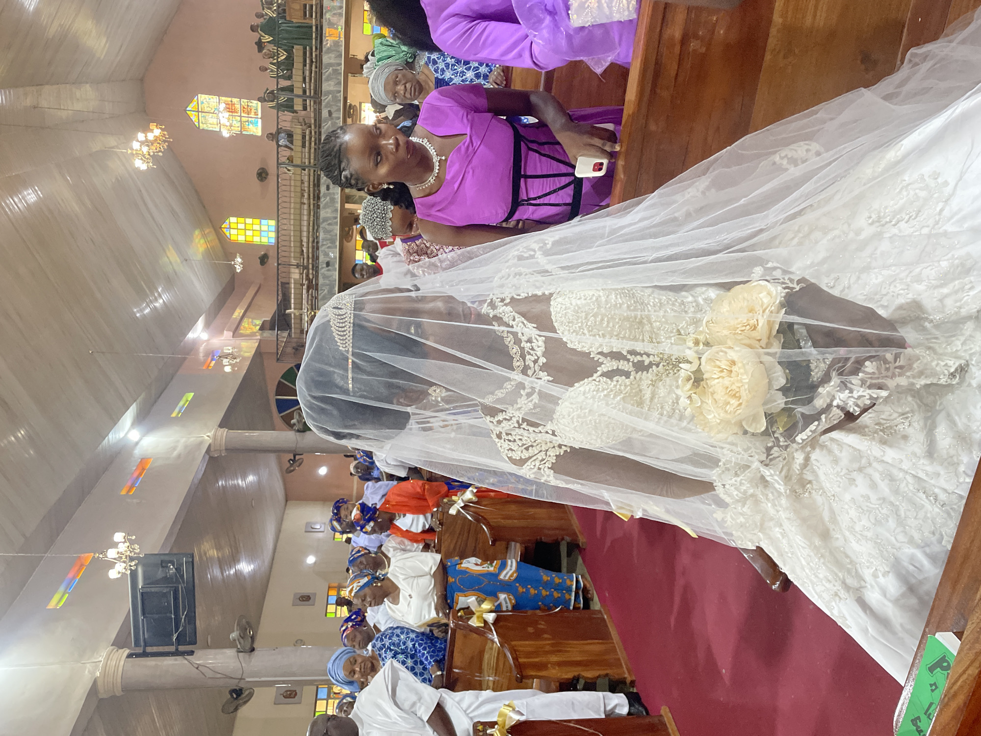 A wedding is taking place in what appears to be a church interior. A bride in a white wedding gown with a veil and a bouquet is the focal point, positioned in the foreground. A woman in a purple dress is seated in the pew beside her, smiling and holding a white smartphone. Numerous people are seated in pews behind the bride, dressed in a variety of colors. The interior features stained glass windows and chandeliers. A red carpet runs along the center aisle, with wooden pews arranged on either side. Based on the mention of Gura Top, Nigeria, it can be inferred that this event is happening there.