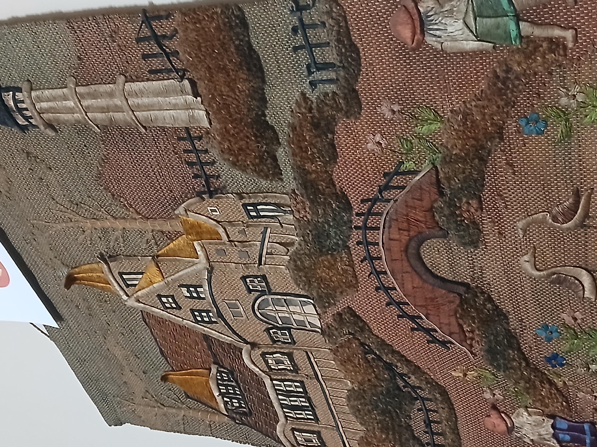 The image depicts a detailed, embroidered artwork of a cityscape. The artwork features buildings with distinctive gabled roofs, a winding path or river with a bridge, and figures of people in the foreground and background. The embroidery uses a variety of colors and textures to create depth and dimension, and the perspective is somewhat stylized. Details include individual windows on the buildings, trees lining the path, and flowers scattered throughout the scene. The image does not contain information regarding the location being Bengaluru, India. The artwork is shown at an oblique angle, possibly displayed on a wall.