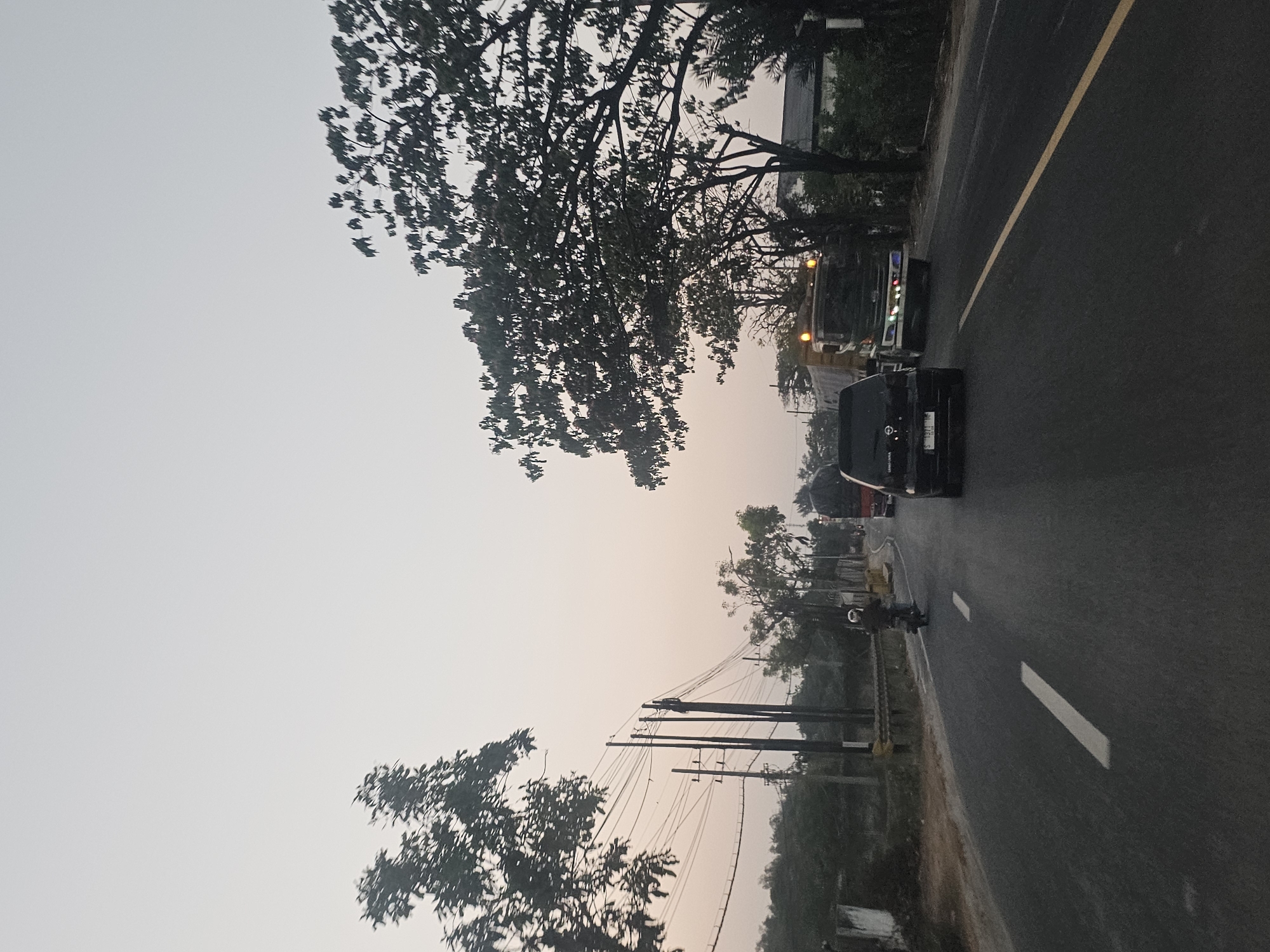The image depicts a road scene in Jenu, Indonesia, at what appears to be dusk or dawn due to the muted, hazy sky and soft lighting. The road is paved and marked with white dashed lines indicating lanes. On the right side of the road, a yellow line separates the lanes.
Several vehicles are visible. In the foreground, a black SUV is traveling on the right side of the road. Further ahead, a large truck with its headlights and additional lights illuminated is also traveling on the right. To the left of the truck, a lighter-colored truck is partially visible. A motorcycle with a rider wearing a helmet is on the left side of the road, moving in the same direction as the other vehicles.
The roadside is lined with trees, some with dense foliage and others appearing sparser. Utility poles with numerous wires are prominent on the left side of the road, suggesting the presence of electricity or telecommunication infrastructure. A low wall or embankment is visible along the left side of the road. In the distance, buildings and more vegetation can be observed. The overall atmosphere is that of a typical road in a semi-urban or rural area during low light conditions.