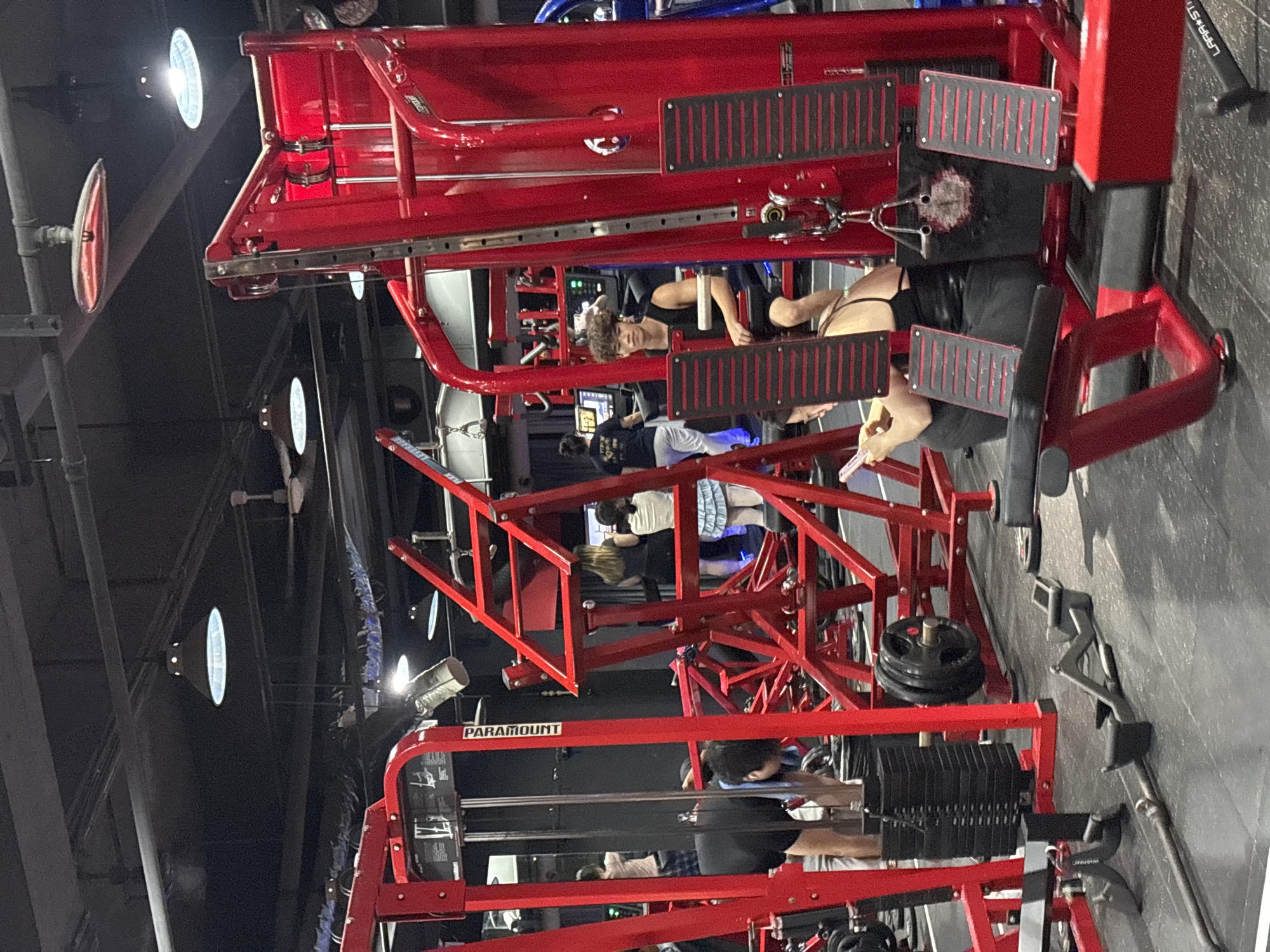 The image shows the interior of a gym. A person is using a red leg press machine with black plates. The "PARAMOUNT" brand is visible on the equipment. Other people are in the background, some using other exercise machines. The location is likely in an urban area, possibly in the Nan'an District, China, based on the prompt. There are overhead lights and a grey floor.