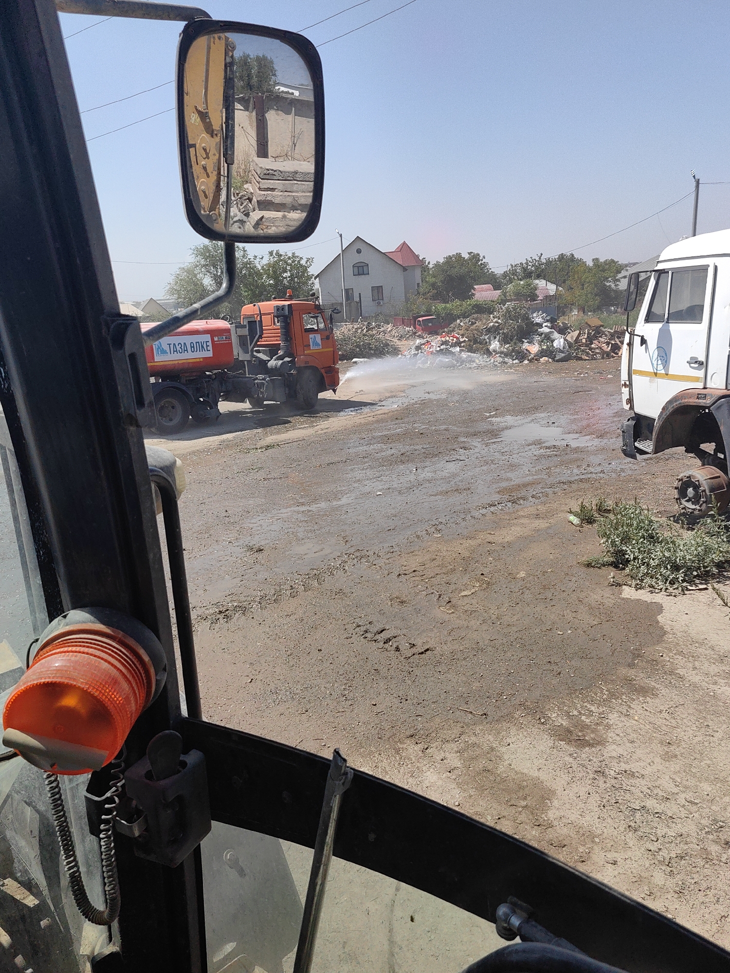 The image, captured from inside a vehicle's cabin, shows an outdoor scene in Shymkent, Kazakhstan. A red water truck, labeled with Cyrillic text that translates to "Clean Water," is spraying water onto a dirt road. The truck has a logo resembling a stylized blue bird. In the background, a large pile of debris and trash is visible, with several buildings and trees beyond it. To the right, a white truck with a missing wheel is parked. The sky is clear and blue, indicating a sunny day. The scene appears to be a street or an open area where cleanup or dust suppression is occurring.