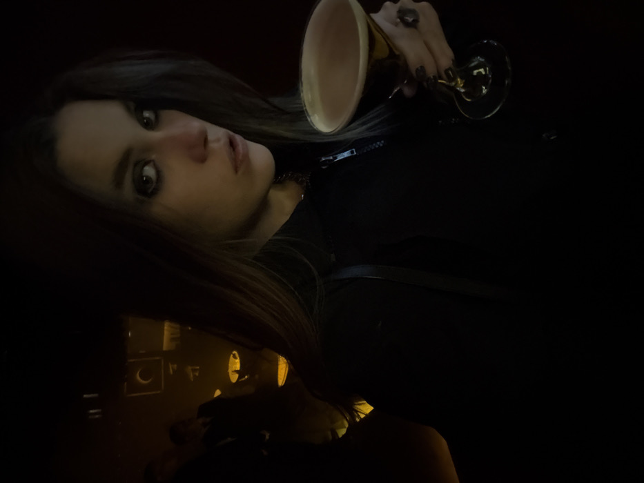 A woman with long brown hair, wearing a black jacket, holds a goblet. She has a gold chain necklace and a ring on her hand. The scene is dimly lit, with the woman's face and the goblet being the most prominent features. A reflective surface below reveals vague illuminated shapes. The woman appears to be looking directly at the viewer.
