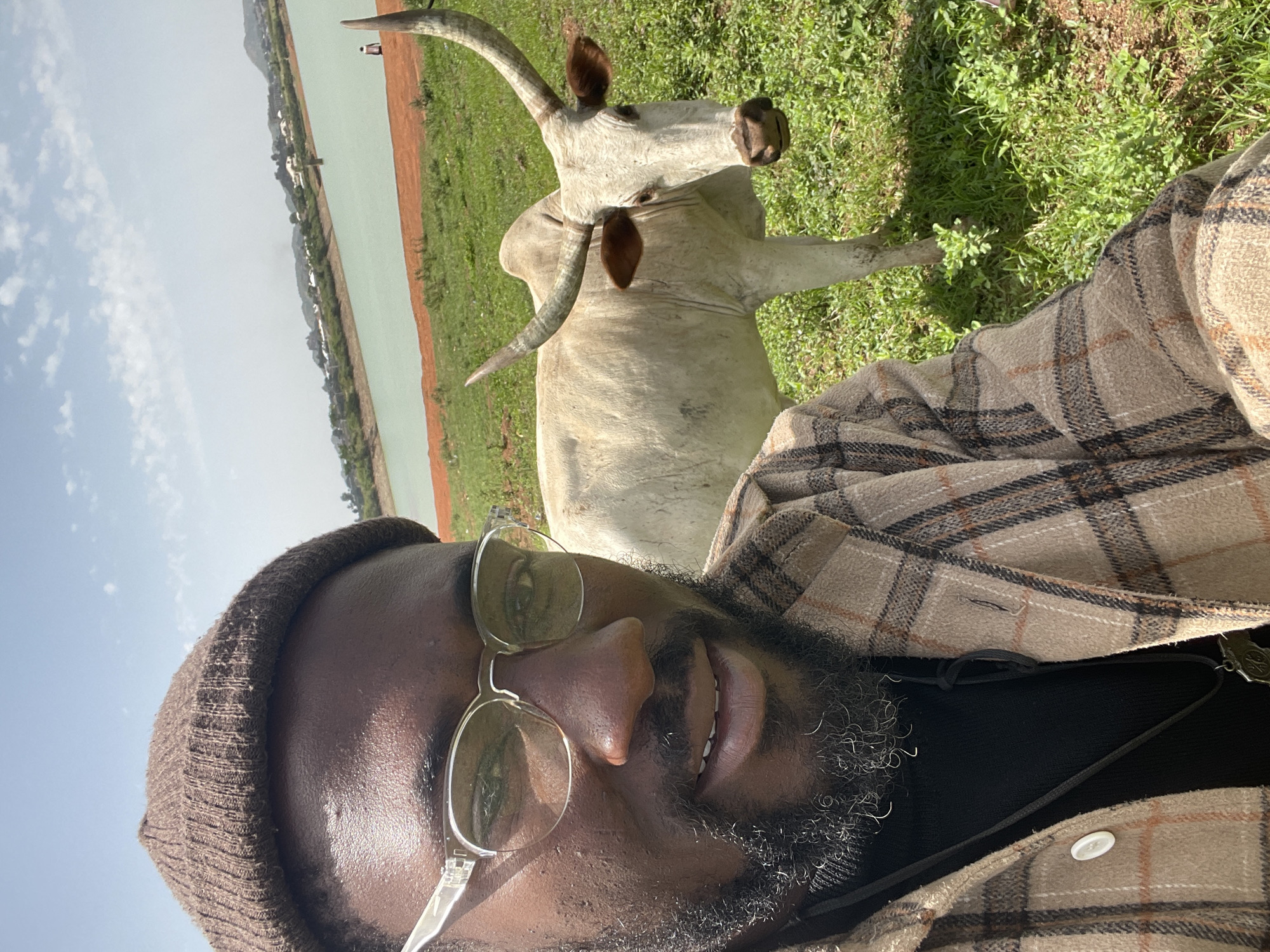A selfie taken by a man in an outdoor environment. He is wearing a brown beanie, clear-framed glasses, and a plaid shirt. He has a beard. Behind him is a white cow with long, curved horns, standing in a grassy area. In the background, there is a body of water with a reddish shoreline. The scene is in Jos, Nigeria.