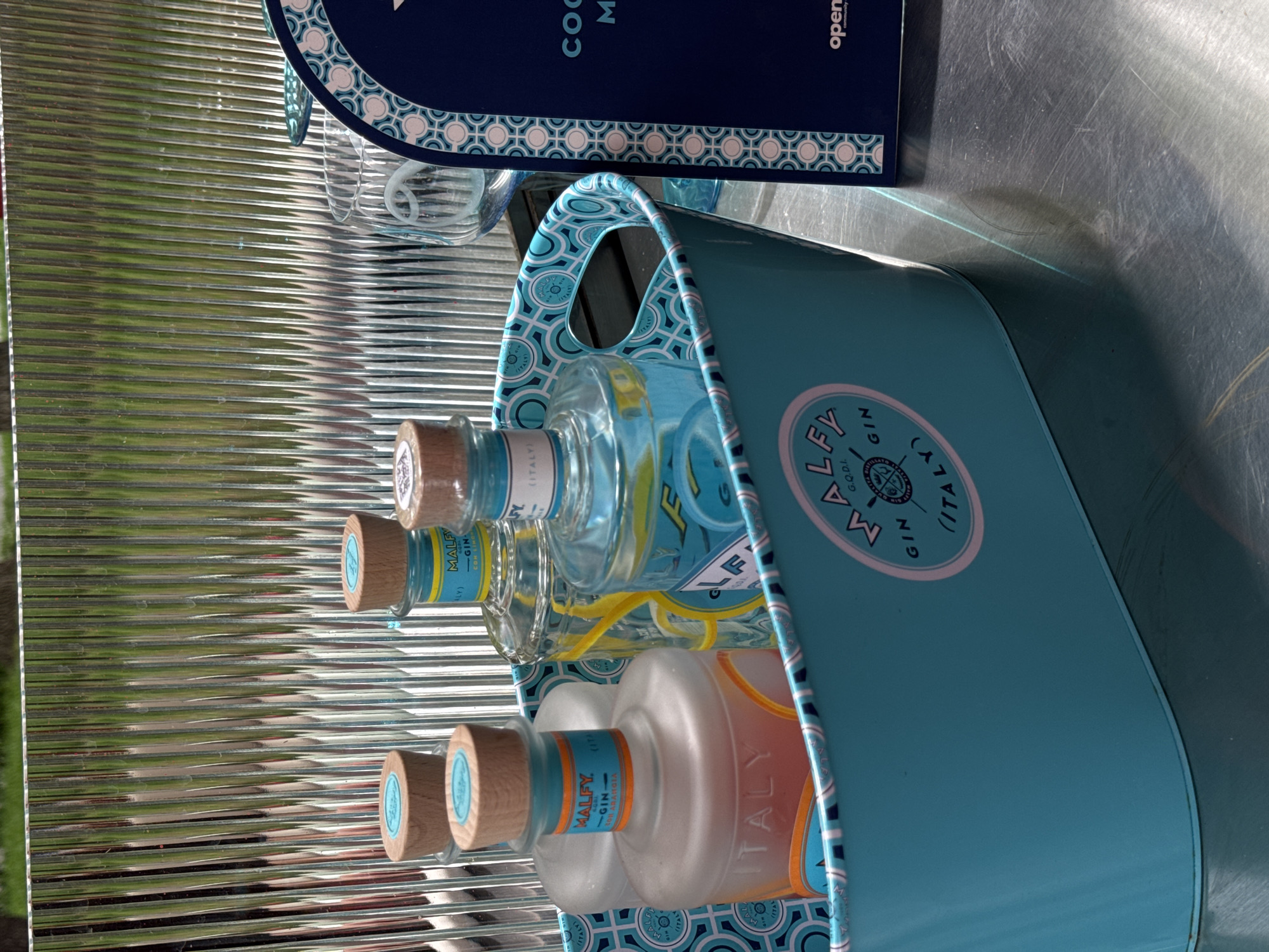 A still-life photograph showcases a blue container filled with gin bottles, likely Malfy Gin given the branding on the bottles and container. The bottles have wooden caps and the gin appears to be clear, with one bottle having an orange hue. The container has a geometric pattern on the trim. The scene is set against a corrugated metal surface, and part of a dark blue object is visible in the upper portion of the frame. The location is unknown, but the request to specify Yerevan, Armenia, suggests it may be there.