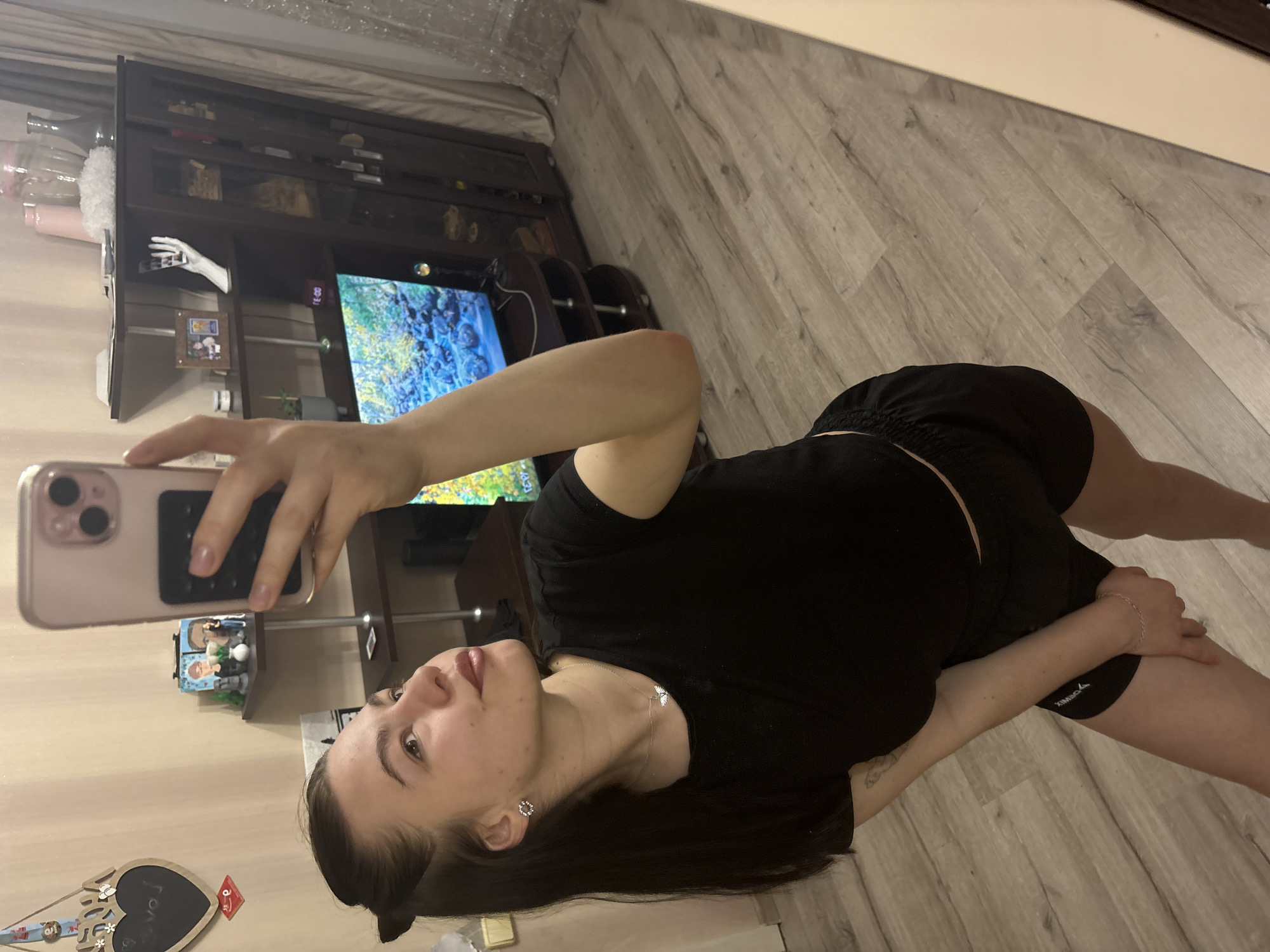 The image depicts a woman taking a selfie in an indoor environment, likely a home in Perm, Russia based on visual cues such as the architectural style. The woman is wearing black workout shorts and a black t-shirt. She is holding an iPhone in her right hand to take the picture, and the phone is in a light pink case. She has dark hair and is wearing a necklace and earrings. The background includes a wooden floor, a TV stand with a TV displaying a colorful scene, and a shelving unit or cabinet with various decorative objects, including a white mannequin hand, and a framed picture. The walls are light-colored and adorned with decorative items. The lighting is soft, suggesting indoor lighting conditions. The image appears to be taken at an angle, tilting towards the right.