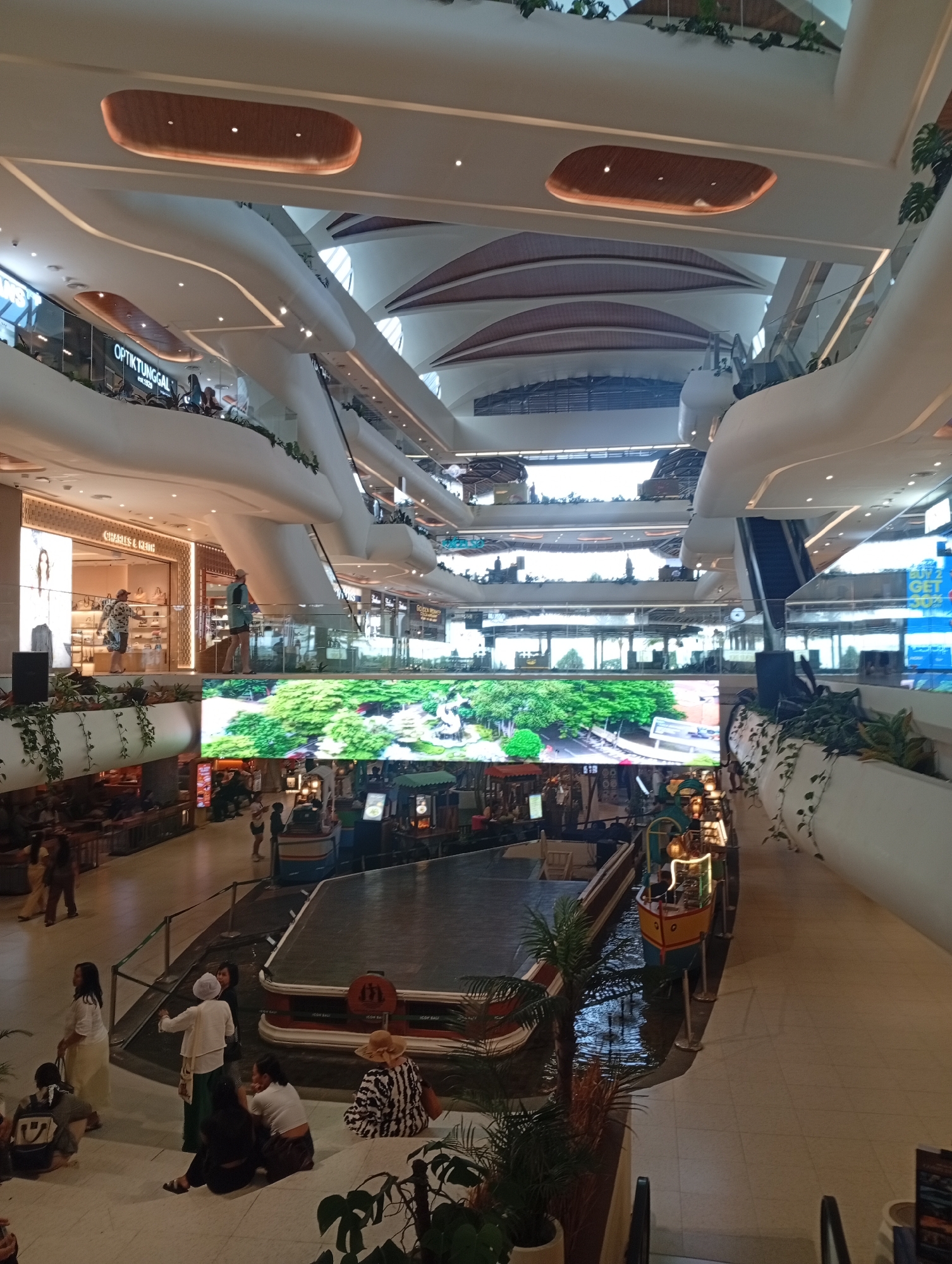 This media file depicts the brightly lit, multi-level interior of a modern shopping mall, likely in Denpasar, Indonesia. The architecture is characterized by elegant, curvilinear white structures, open spaces, and numerous escalators connecting the various floors. Wooden accents, shaped somewhat like inverted boats, adorn the ceilings, interspersed with recessed spotlights. Green foliage is integrated into the design, hanging from ledges and planters.
A prominent feature on the ground floor is a canal-like water body, within which several colorful, decorative boats, reminiscent of traditional vessels, are moored. Above this, a large LED screen displays a vibrant, lush green landscape, creating a striking visual centerpiece.
Many people are visible throughout the mall. On the lower level, a group of individuals is seen sitting casually on wide steps near the water feature, observing the surroundings. Other people are walking along the paths or gathered near the shops. On the upper levels, shoppers stroll past retail outlets.
Visible text includes shop names like "OPTIKTUNGGAL est.1929" and "CHARLES & KEITH." The mall is well-lit, suggesting active business hours, and the atmosphere appears relaxed and leisurely. The combination of modern design, integrated greenery, and the unique water feature with boats suggests a blend of commerce and thematic entertainment.