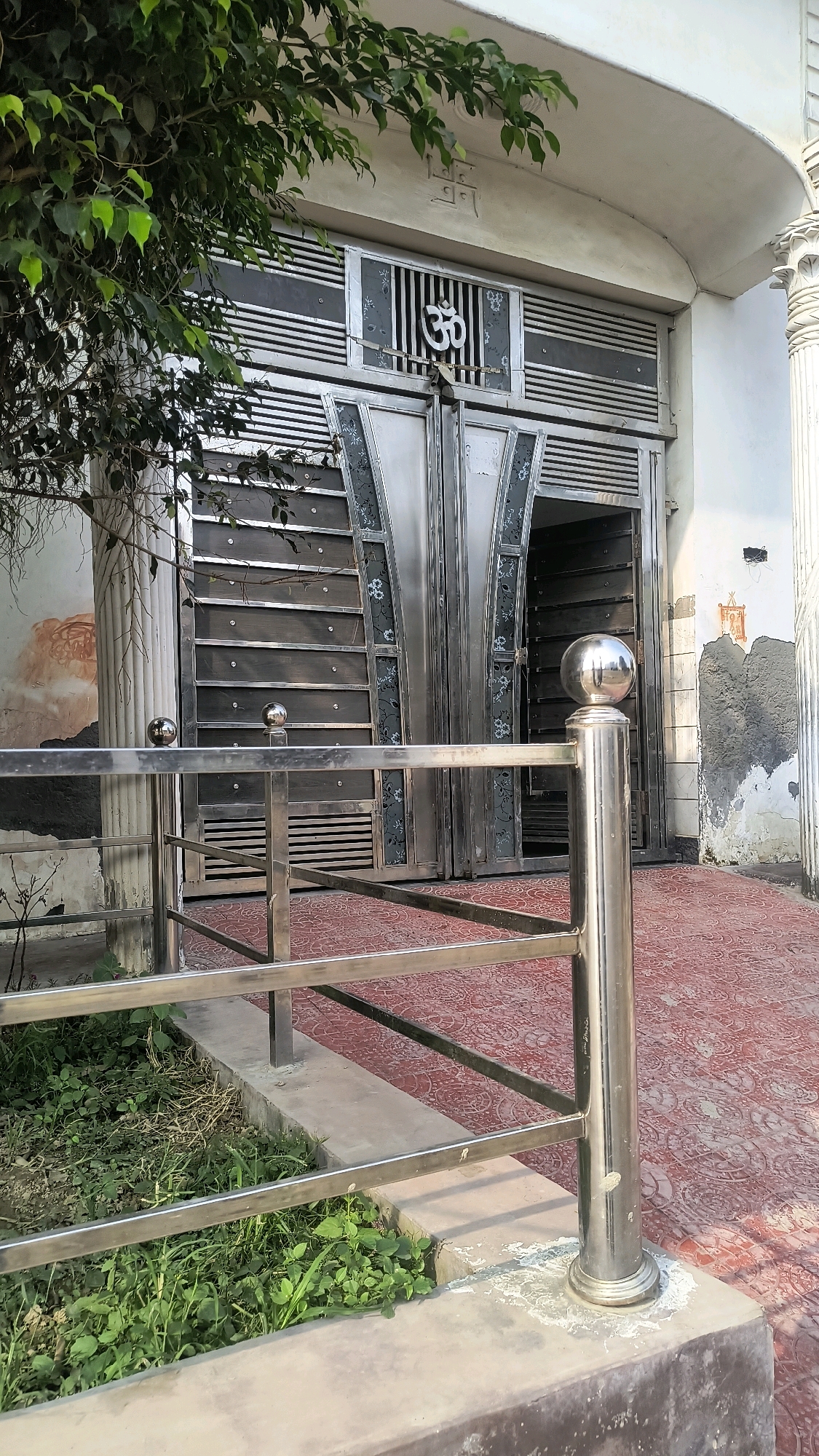 The image shows a metallic gate with an Om symbol. The gate is decorated with a floral pattern and is flanked by fluted columns. The ground is tiled in red with a patterned design. A metallic railing is in the foreground, with lush green plants growing behind it. The scene appears to be located in Loni, India.