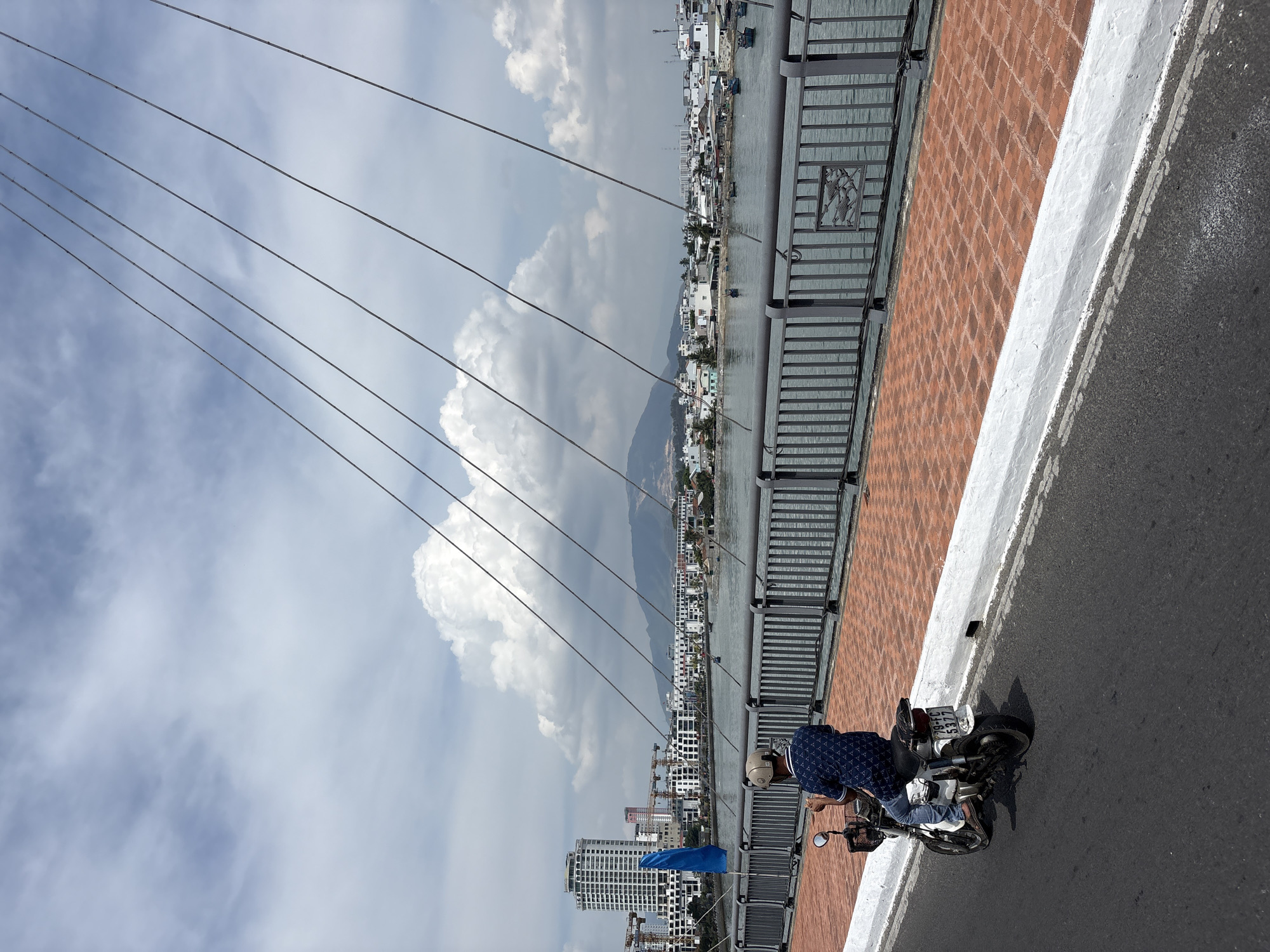 A man on a scooter is on a bridge in Nha Trang, Vietnam. The bridge has a brick walkway with a metal guardrail and suspension cables. Across the water are buildings and a mountain. The sky is cloudy.