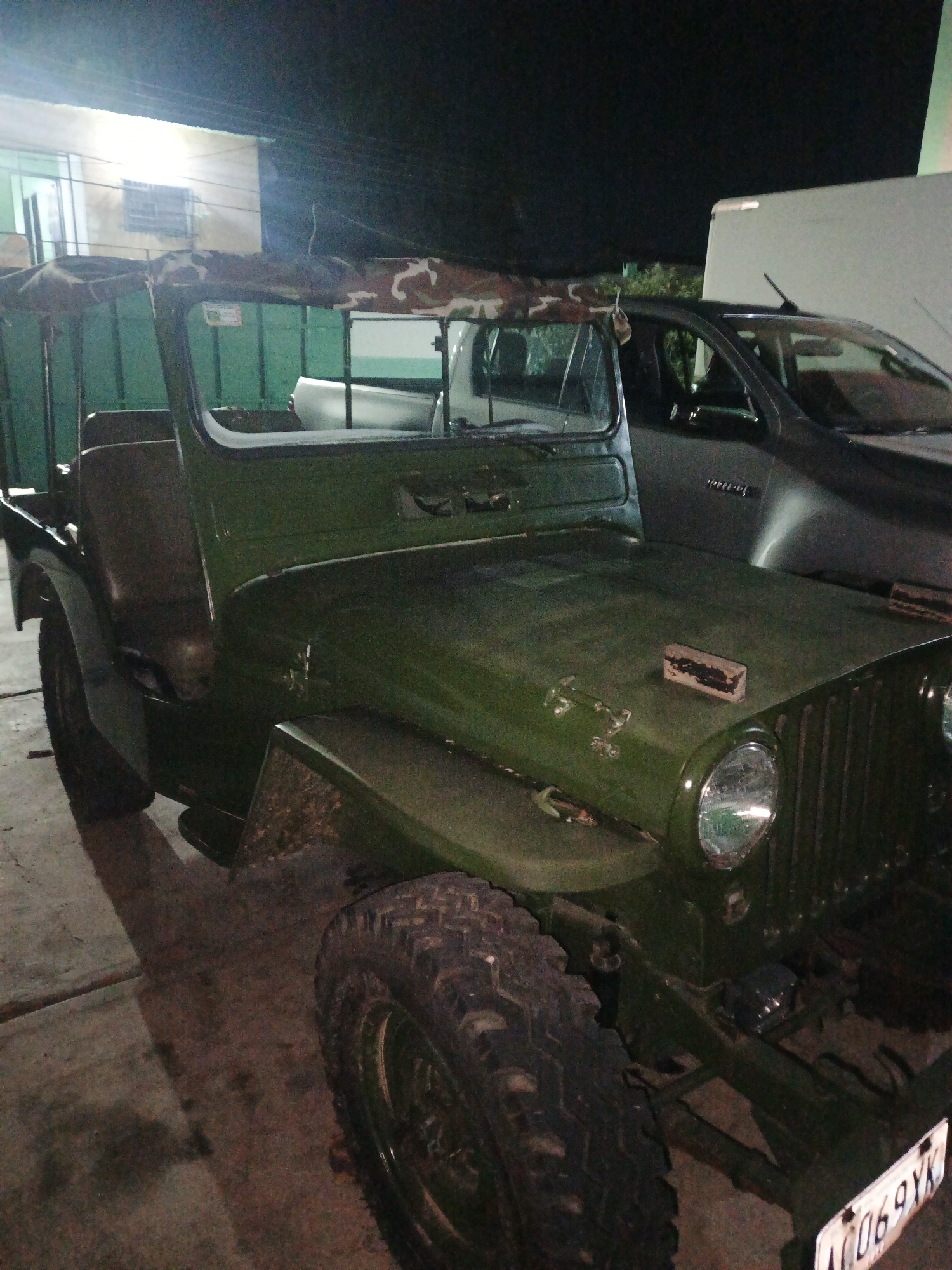 The image shows a dark green, vintage Willys Jeep parked outdoors at night, possibly in the city of Tinaquillo, Venezuela, judging from the license plate visible on the front bumper. The vehicle has a camouflage patterned soft top. A dark grey modern truck is parked behind the Jeep. The lighting is poor, with an overhead light source visible near what appears to be a building with green doors.