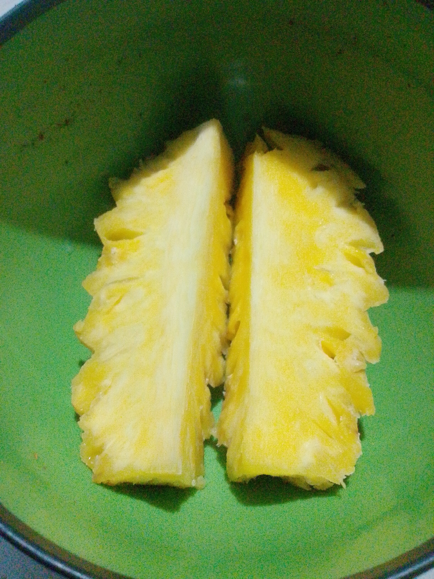 Two pieces of pineapple are arranged in the center of a green bowl. The pineapple pieces are cut lengthwise and appear to be fresh. The bowl is round and has a slight sheen to its surface. The background is a solid green, likely the interior of the bowl. There are no people visible in the image. The scene is likely a kitchen or dining area. This image has no specific details relating to the city of Bandar Lampung, Indonesia.