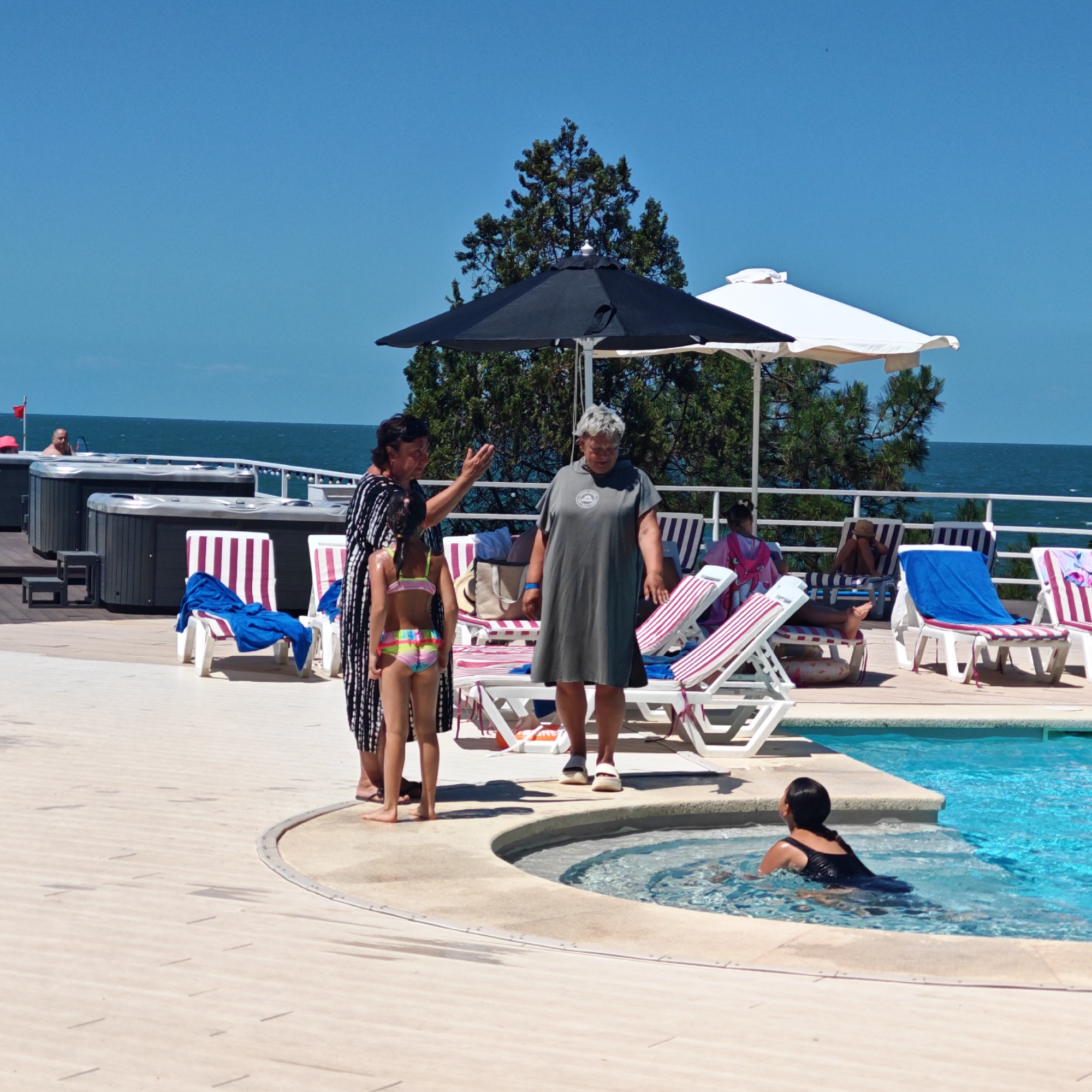 This image depicts a sunny outdoor scene, likely at a resort or spa, given the presence of hot tubs, lounge chairs, and a swimming pool. The setting appears to be a paved area next to a body of water, possibly the Black Sea, as indicated by the horizon line and the presence of umbrellas and trees typical of coastal regions. The time of day is midday, suggested by the bright sunlight and distinct shadows.
In the foreground, several people are visible. A woman in a black and white patterned dress is speaking to a young girl wearing a colorful bikini. Another woman, dressed in a grey, knee-length dress, stands nearby. To the right, a person in a black swimsuit is partially submerged in the clear blue water of a swimming pool. In the background, two large hot tubs are situated on a wooden deck, with a man visible in one of them. Several lounge chairs with striped cushions are arranged around the pool area.
The overall atmosphere is relaxed and leisurely, consistent with a vacation or leisure setting. The people do not display overt emotions, but their attire and the environment suggest a warm, pleasant day.
There is no visible text in the image.