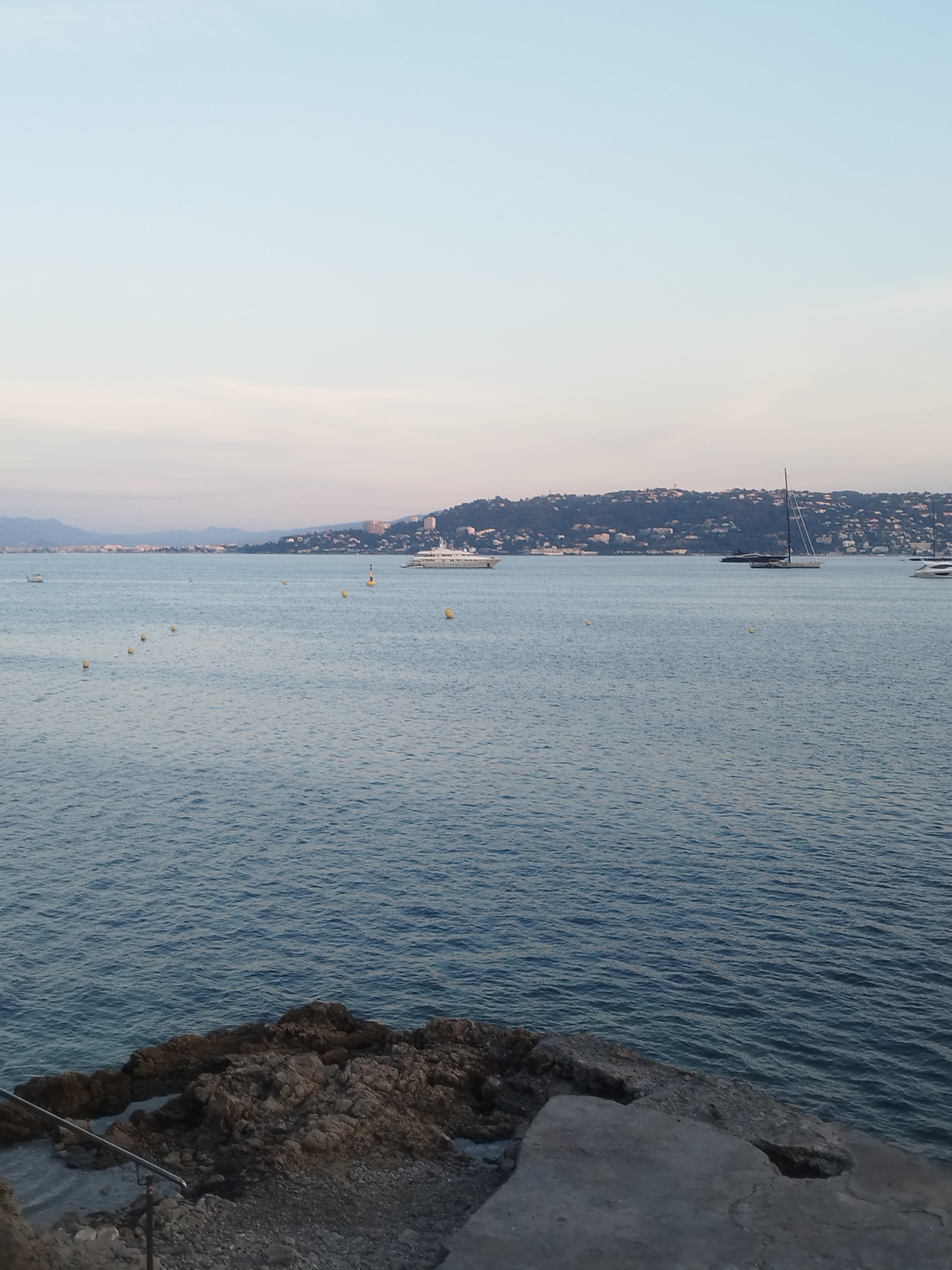 The image captures a serene coastal view in Antibes, France, likely from a viewpoint above the water. The foreground features a rocky shoreline with some concrete structures and a metal railing, suggesting a public walkway or an observation point. The sea, a deep blue, stretches to the horizon, dotted with several yellow buoys. In the mid-ground, a large white yacht is anchored, and further out, a dark sailboat with tall masts is visible. The background displays a hillside town with buildings nestled amongst lush greenery, characteristic of the Antibes coastline. The sky is a soft gradient of pale blue and pink, indicating either early morning or late evening. The overall atmosphere is peaceful and tranquil, highlighting the beauty of the Mediterranean coast.