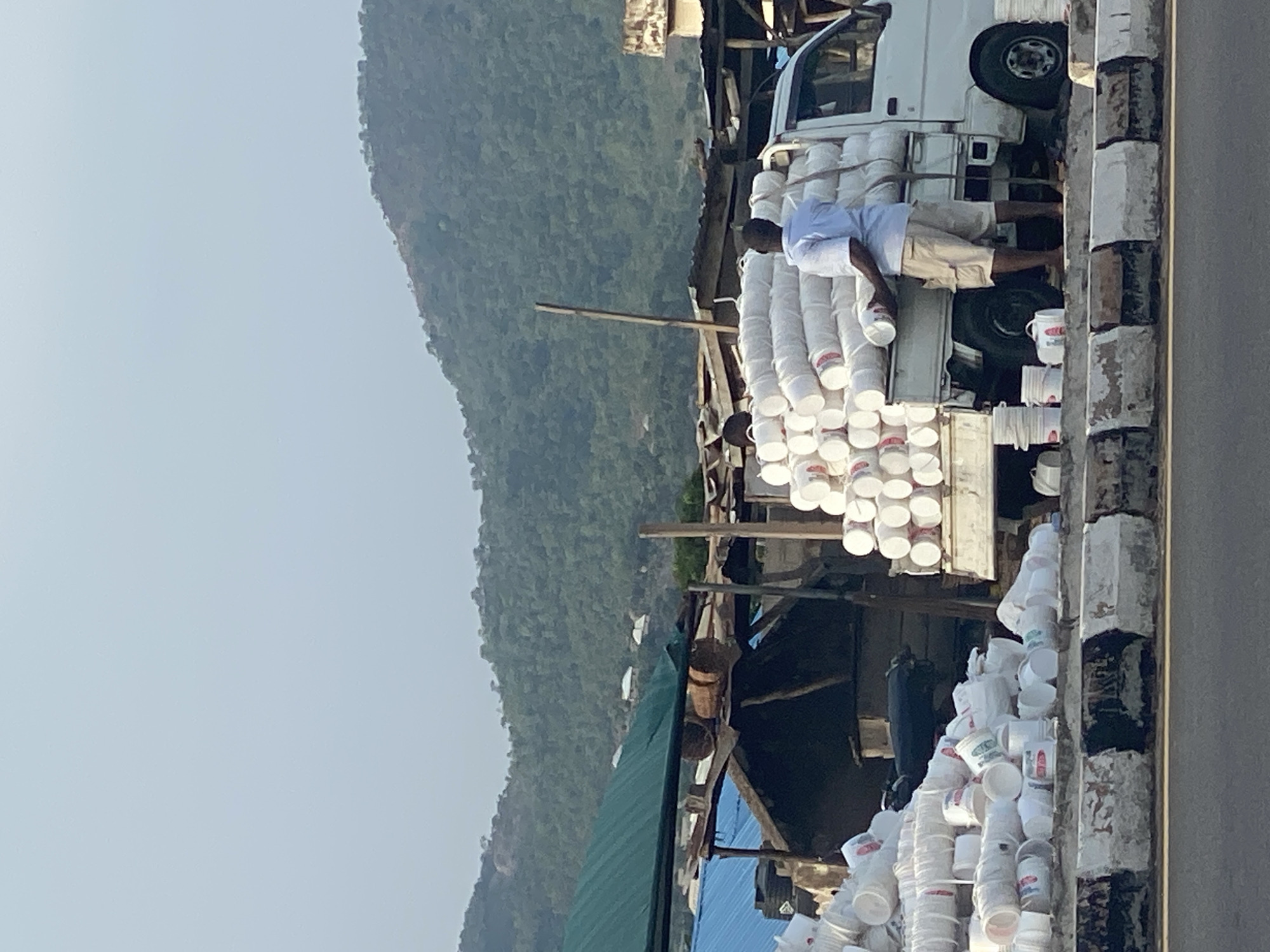 This image, set in Adankolo, Nigeria, depicts an outdoor scene featuring commercial activity. A white pickup truck, positioned centrally, is heavily loaded with numerous white plastic buckets or containers, stacked high on its bed. A man, wearing a light blue short-sleeved shirt and light-colored shorts, stands on the truck bed, appearing to be arranging, loading, or securing these buckets. Additional white buckets are visible on the ground next to the truck.
To the left of the truck, there is a simple structure with wooden supports and a green tarp serving as a roof, possibly indicating a market stall or temporary shelter. A paved road or path with a concrete curb or barrier runs along the right side of the frame, adjacent to the truck. The background is dominated by large, verdant hills or mountains covered in dense vegetation, rising beneath a bright, somewhat hazy sky.
The scene takes place during daylight hours, characterized by bright, even illumination. The activity suggests a busy commercial operation involving the transport or distribution of goods, likely contained within the white buckets. While some buckets show faint colored markings or labels, no specific text is clearly legible. The overall impression is one of industrious roadside commerce in a naturally scenic, hilly environment.