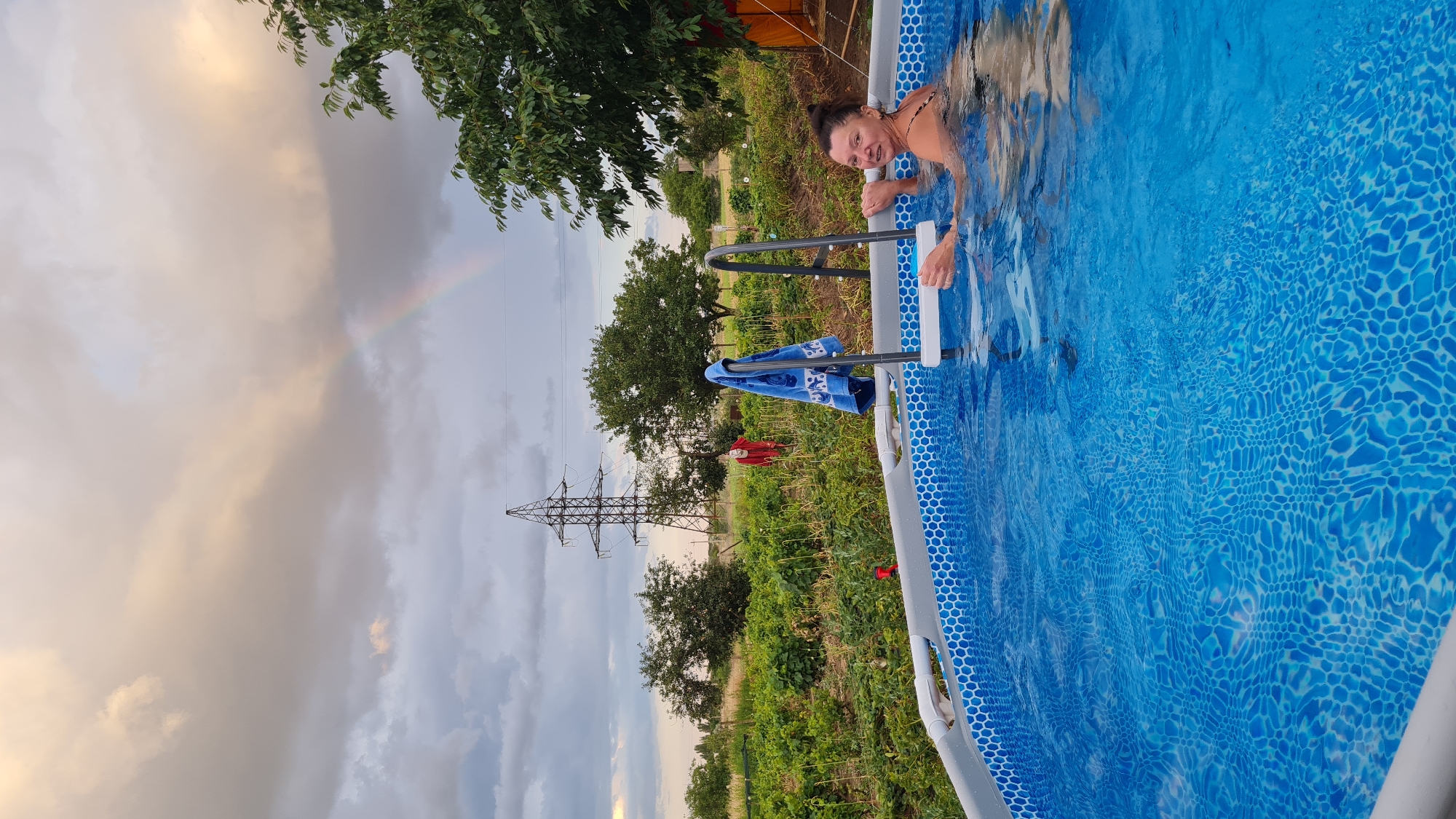 The image depicts an outdoor swimming pool with a woman in it, located in Salhany, Ukraine. The woman is partially submerged in the blue water, wearing a black and white patterned swimsuit, and appears to be smiling at the camera. A blue towel with a white pattern is draped over the pool ladder. The pool is surrounded by a garden with lush green plants. In the background, a tall electrical pylon stands against a cloudy sky, with a faint rainbow visible in the distance. The overall scene suggests a rural or suburban setting.
