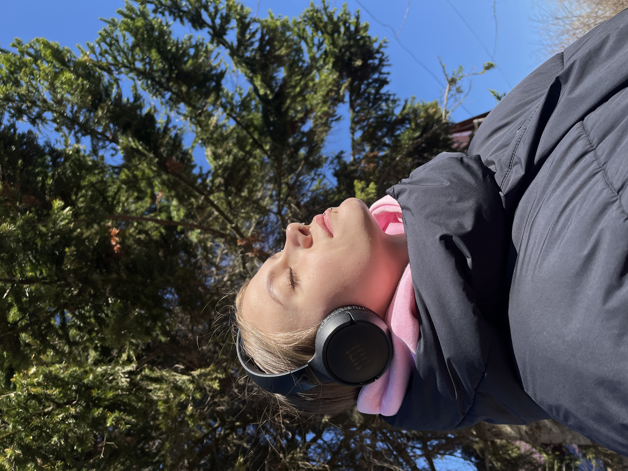 A young person is outdoors with their head tilted back. The person is wearing headphones, a pink hooded shirt, and a black jacket. The background includes a large tree and a blue sky. The city Фоки, Russia, is not visible or discernible in the image.