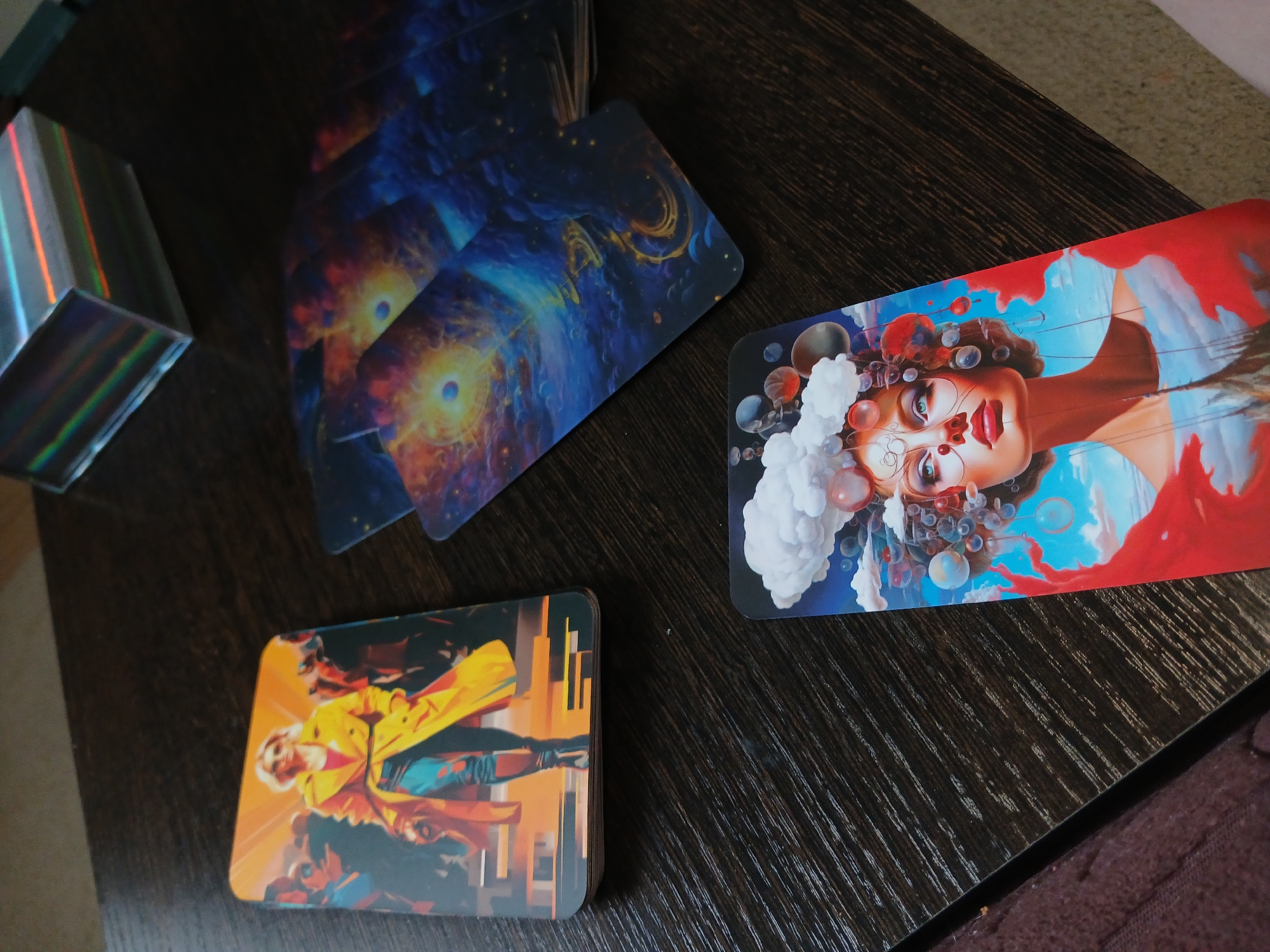 A high-angle shot displays three tarot cards and a holographic box on a dark, wooden surface. The card in the foreground on the bottom right features a close-up of a woman's face, adorned with clouds, bubbles, and abstract shapes, with a red scarf draped around her shoulders. To the left, a second card shows a woman in a bright yellow coat and sunglasses, walking amidst a crowd. In the middle right, a stack of cards displays a cosmic scene with swirling galaxies and celestial bodies. A small, holographic box with rainbow-colored edges sits in the upper right corner. The scene appears to be indoors, possibly on a table. There is no discernible connection to the city of Yekaterinburg, Russia.