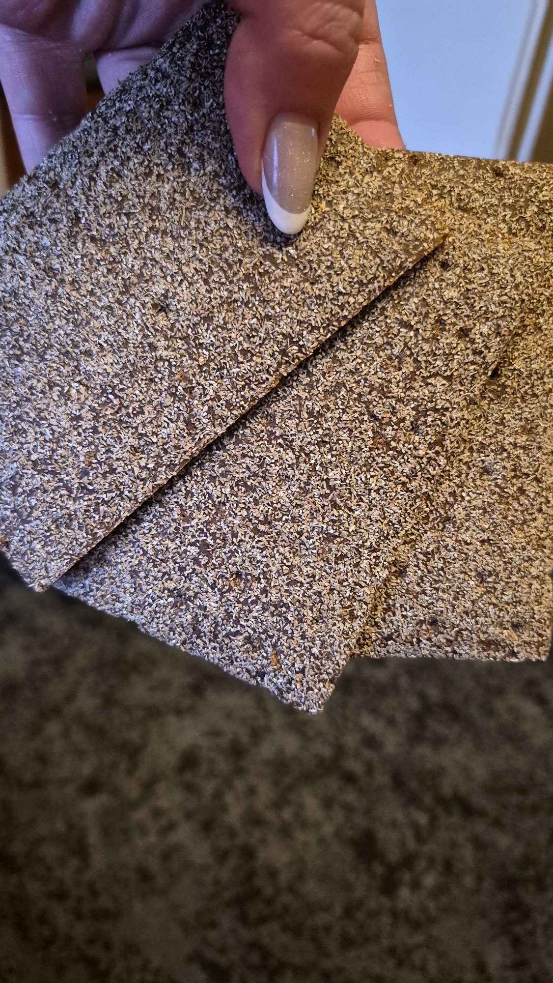 This is a close-up, overhead shot of a person's hand holding a piece of dark brown roofing material with a granular surface. The material is folded, creating a diagonal line across the image. The person's fingernail, painted with a French manicure and glitter, is visible near the top right corner, touching the roofing material. The background consists of a dark, textured surface, possibly carpet or fabric. There is no information available about the location in Krauja, Latvia.