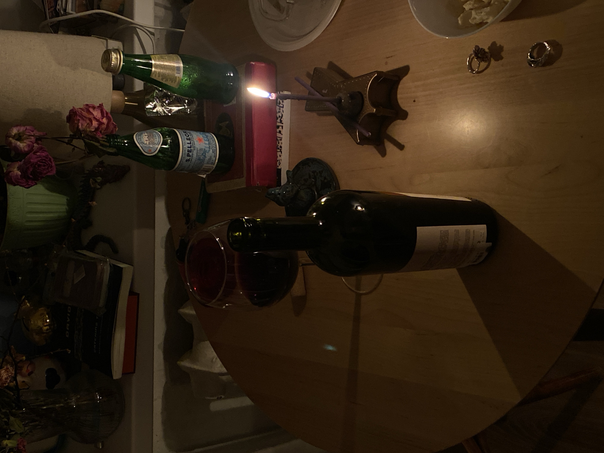 The image depicts a tabletop scene, likely in Заволжье, Russia. Objects include a wine bottle, a glass of red wine, two bottles of mineral water (one San Pellegrino), a candle in a holder, a book, a small vase of dead roses, and two rings. A small bowl contains food items. The lighting is dim, and shadows are prominent.