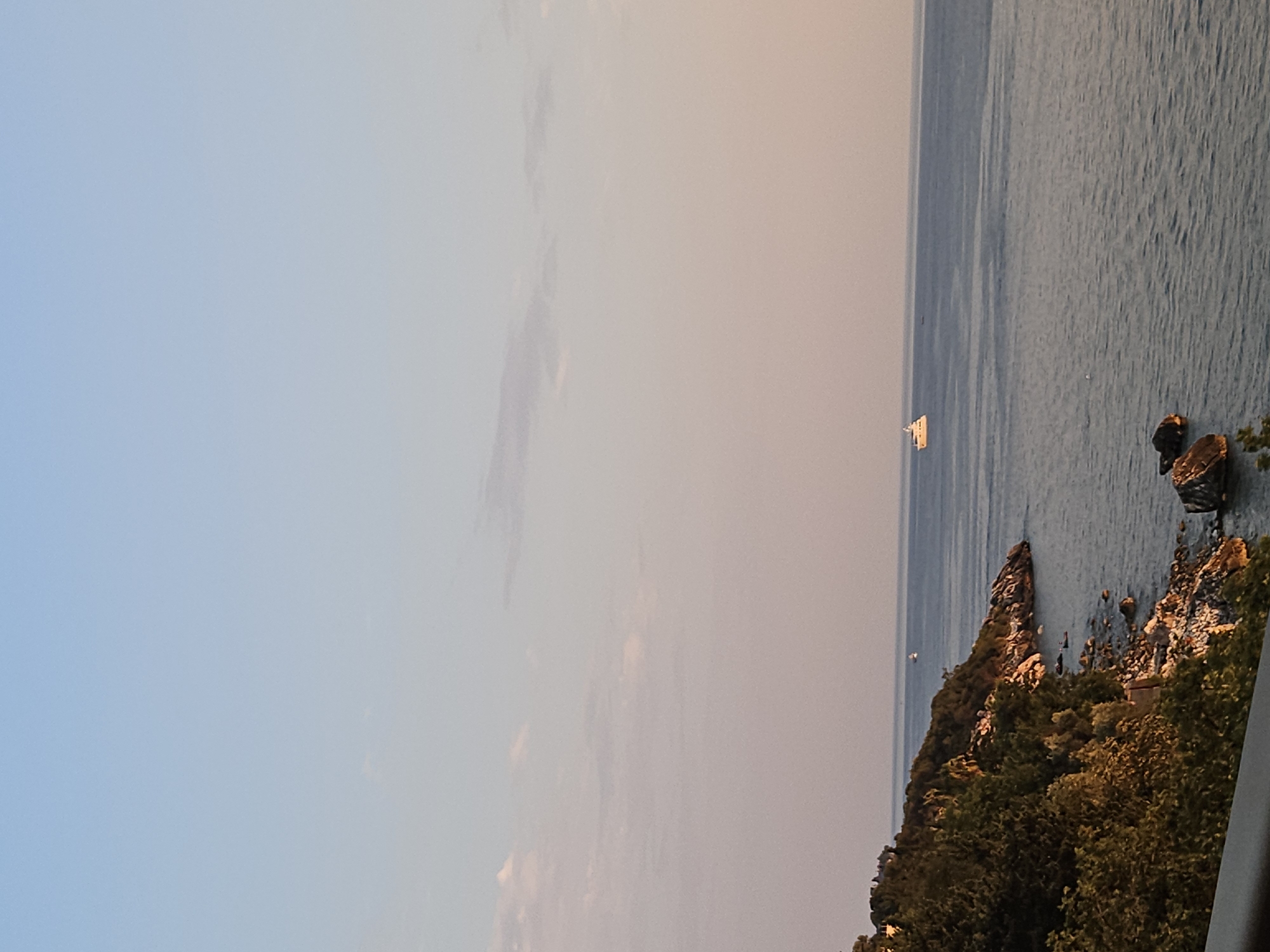 The image shows a body of water with a rocky coastline covered in lush green trees. The sky is a pale blue with some wispy clouds. In the distance, a white yacht is sailing on the water. The sun appears to be low on the horizon, casting a warm glow on the scene. The overall impression is one of peaceful tranquility.