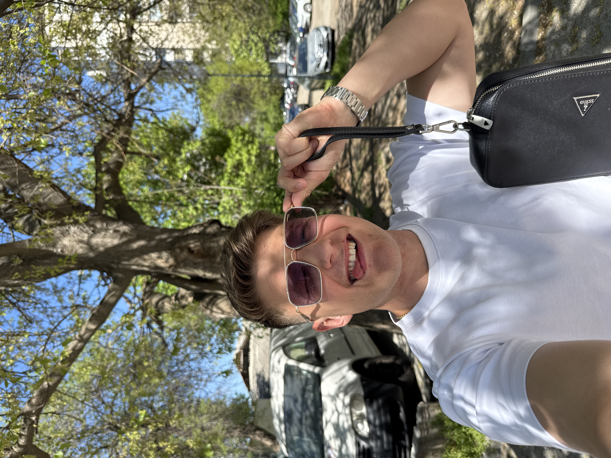 A man stands outdoors, holding a pair of sunglasses and wearing a white t-shirt and a watch. He also holds a black Guess purse. The background includes trees with green leaves, a blue sky, and parked cars. A white van is parked nearby. This photo may have been taken in Plovdiv, Bulgaria.