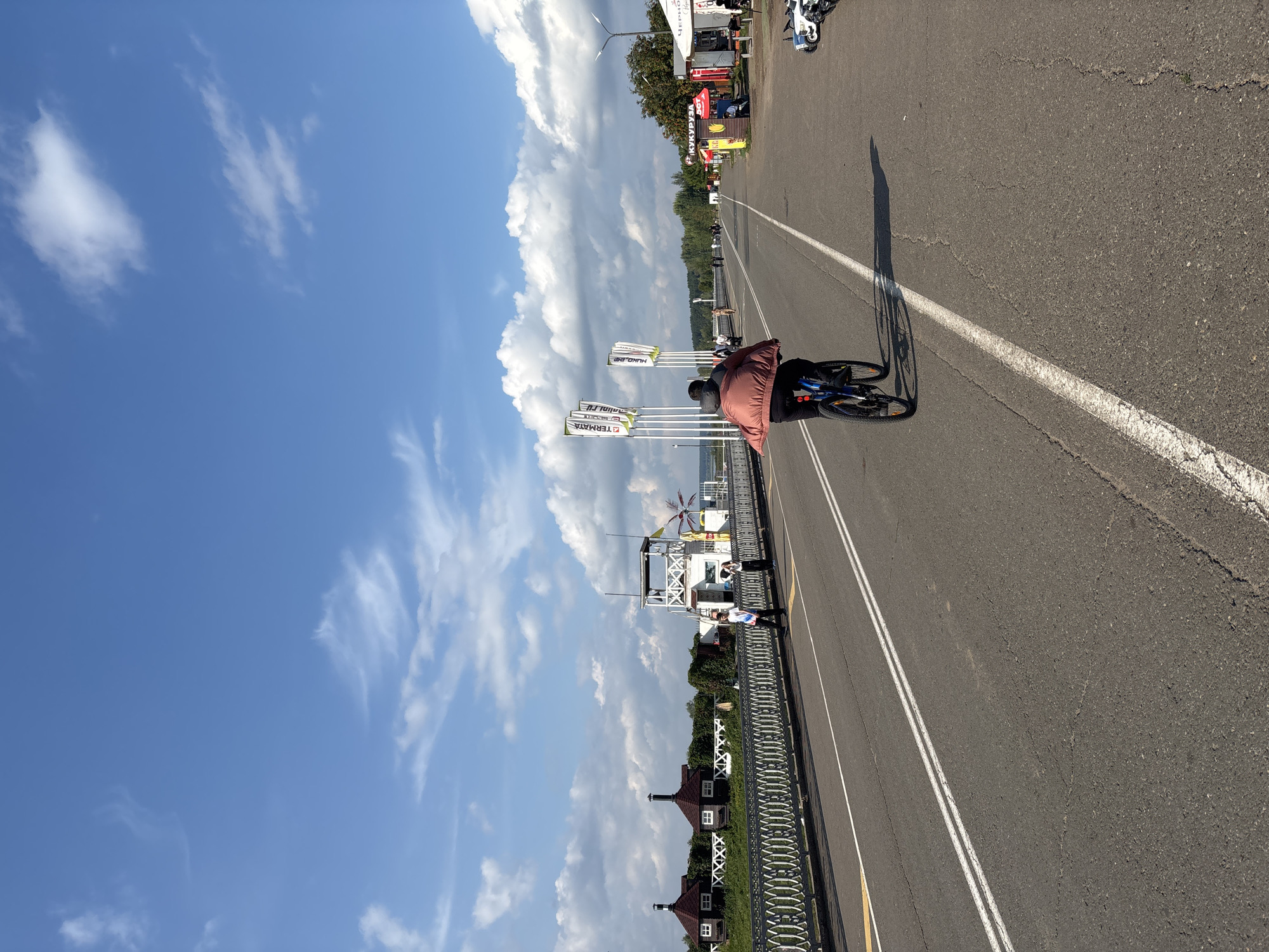 The image depicts a street scene on a sunny day with a blue sky and scattered white clouds. In the foreground, a person rides a bicycle on an asphalt road marked with white lines. The cyclist is wearing dark pants and a reddish-brown jacket. The road appears to be a bridge or elevated causeway, with a decorative metal railing on the left. Beyond the railing, there are several buildings with peaked roofs and chimneys, as well as green foliage. Further down the road on the right side, there are several small vendor stalls and parked vehicles, including what appear to be electric scooters. In the distance, trees line the horizon. The overall scene suggests a public area, possibly a promenade or a section of a city frequented by pedestrians and cyclists. The location is the embankment of the Kama River in Naberezhnye Chelny, Russia, a popular recreational area.