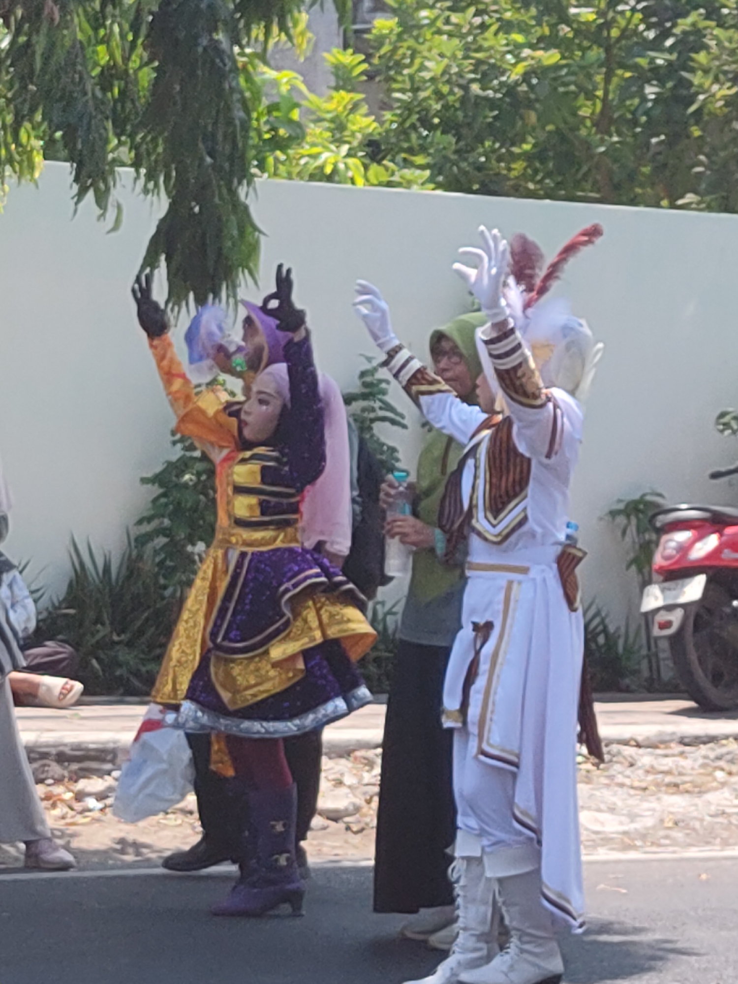 In the city of Ngasem, Indonesia, a group of performers in elaborate costumes are seen standing on a paved street. The central figure on the right wears a white uniform with gold and brown trim, a white feathered headdress, and white gloves, holding one hand raised. To their left, a person in a green hijab stands between them and another performer on the far left. This performer is dressed in a vibrant orange and yellow outfit with a purple, glittery skirt and purple boots, also with a raised hand. In the background, a white wall, lush green foliage, and a red motorcycle parked on the side of the road are visible. The ground is a mix of asphalt and gravel, with scattered leaves and debris.