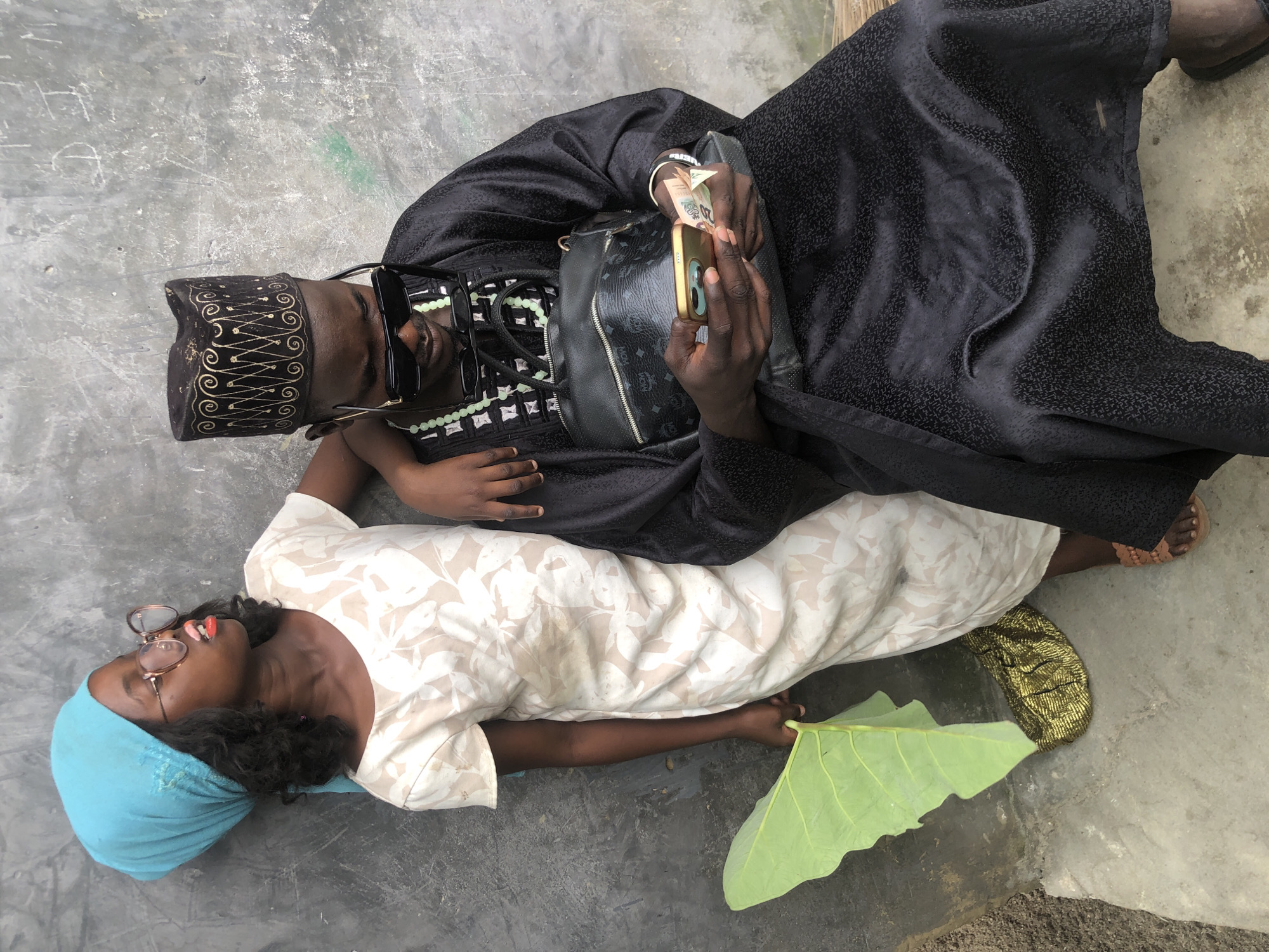 The image depicts a young girl and a young man in what appears to be a gritty urban environment, possibly a street or alleyway in Ado, Nigeria. The girl, wearing a blue headscarf and a light-colored dress with a floral pattern, leans against a rough, grey concrete wall. She has large, round glasses and her mouth is slightly open with red lipstick visible. She is holding a large, vibrant green leaf.
The young man, seated next to her, is dressed in traditional black attire, including a kufi cap adorned with gold embroidery. He wears dark sunglasses and a necklace of light green beads. He is engrossed in his mobile phone, holding it in his right hand while his left hand holds some banknotes. A black handbag with a repeating pattern is slung over his shoulder. A small, gold, textured bag rests on the ground near the girl's feet, and the man is wearing sandals. The overall setting suggests a casual, possibly impromptu, moment captured outdoors, with the textured concrete wall and ground indicating a weathered, unpolished urban landscape.