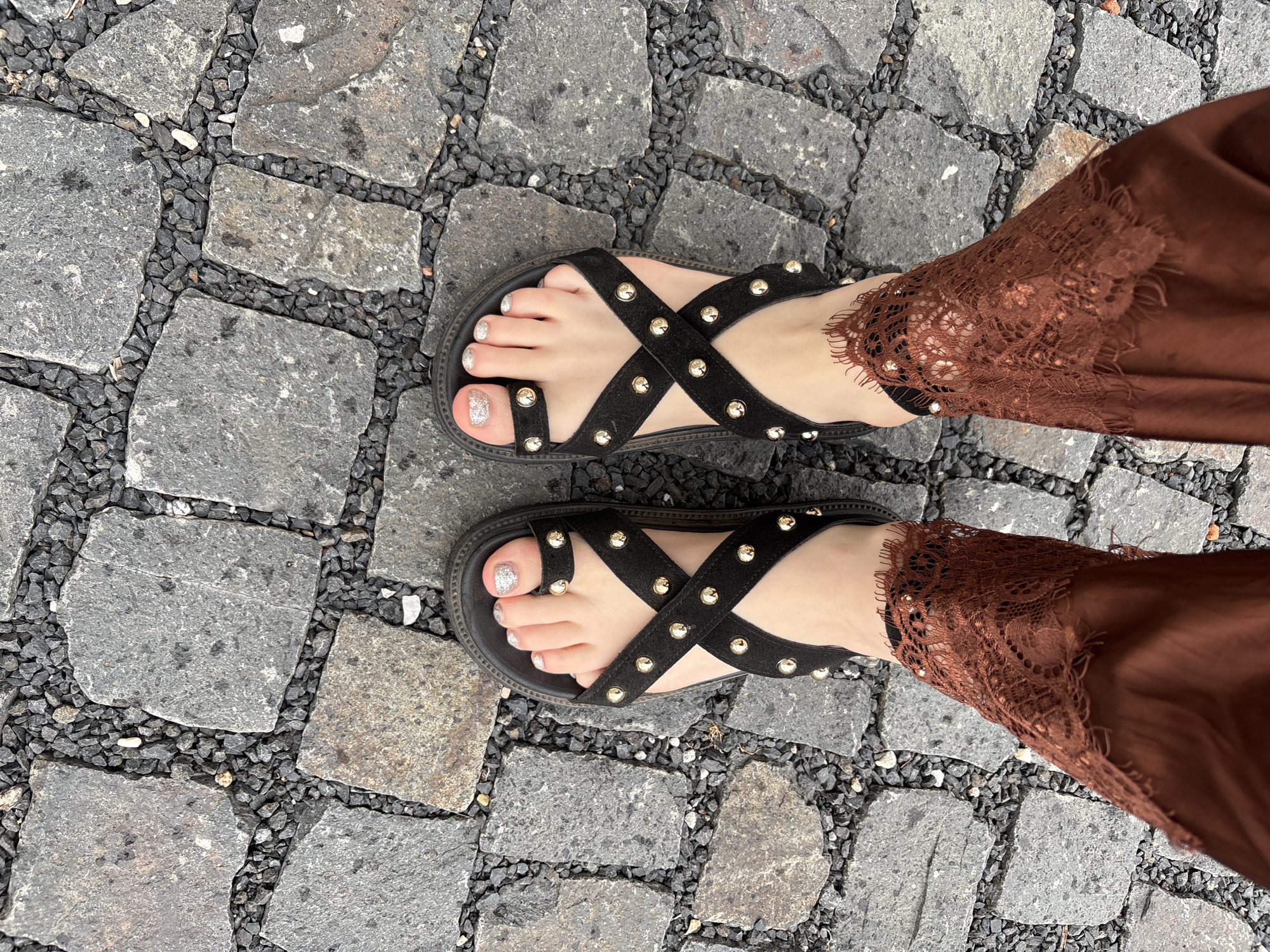 This is a high-angle, medium shot of a person's feet wearing black platform sandals adorned with gold studs. The sandals have crisscrossed straps and are decorated with glittery nail polish on the toenails. The person is also wearing brown lace-trimmed pants that are sheer in places. The feet are positioned on a cobblestone pathway with small dark gray gravel in between the stones. The lighting is bright and natural, suggesting an outdoor setting. The scene appears to be a street or walkway in an urban environment, though the specific city is unknown.