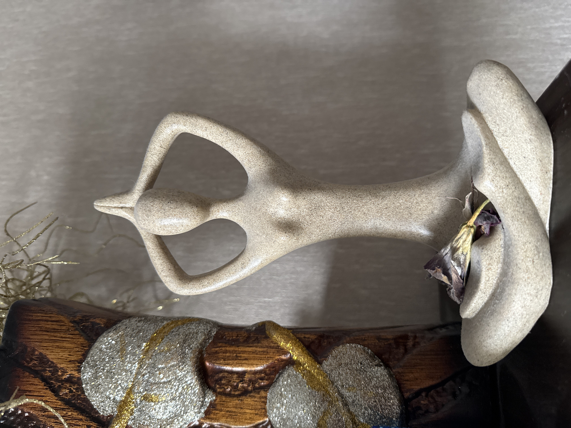 A close-up, frontal view captures a sand-colored, stylized statue of a person in a yoga pose. The statue is positioned on a dark surface, likely a shelf or table, in front of a textured, light-colored wall. To the left of the statue, a portion of a vase or decorative object is visible. This object is dark brown with a textured surface and features a shimmering design of two large, leaf-like shapes outlined in gold and filled with silver glitter. A dried, dark purple flower rests on the base of the yoga statue. The lighting suggests an indoor setting. The image does not provide any visual cues to indicate its location in Bishkek City, Kyrgyzstan.
