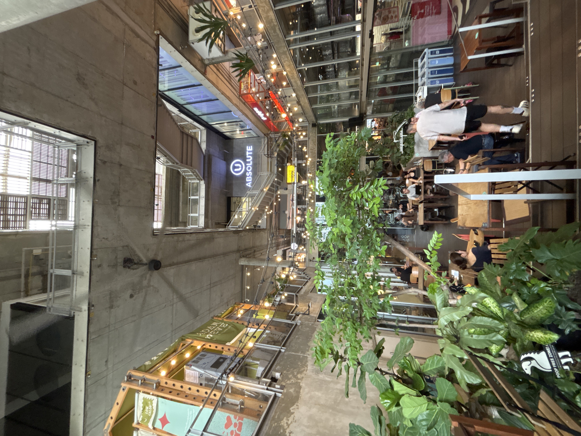 This appears to be an interior view of an open-air commercial space, likely in Bangkok, Thailand. The dominant architectural feature is a raw concrete structure with multiple levels. String lights are suspended throughout the space, creating a casual atmosphere. Various shops or stalls are set up on different levels, showcasing merchandise. There is a sign for "ABSOLUTE" in a prominent location. People are dining, walking, and interacting with the environment. There are various plants in the foreground.