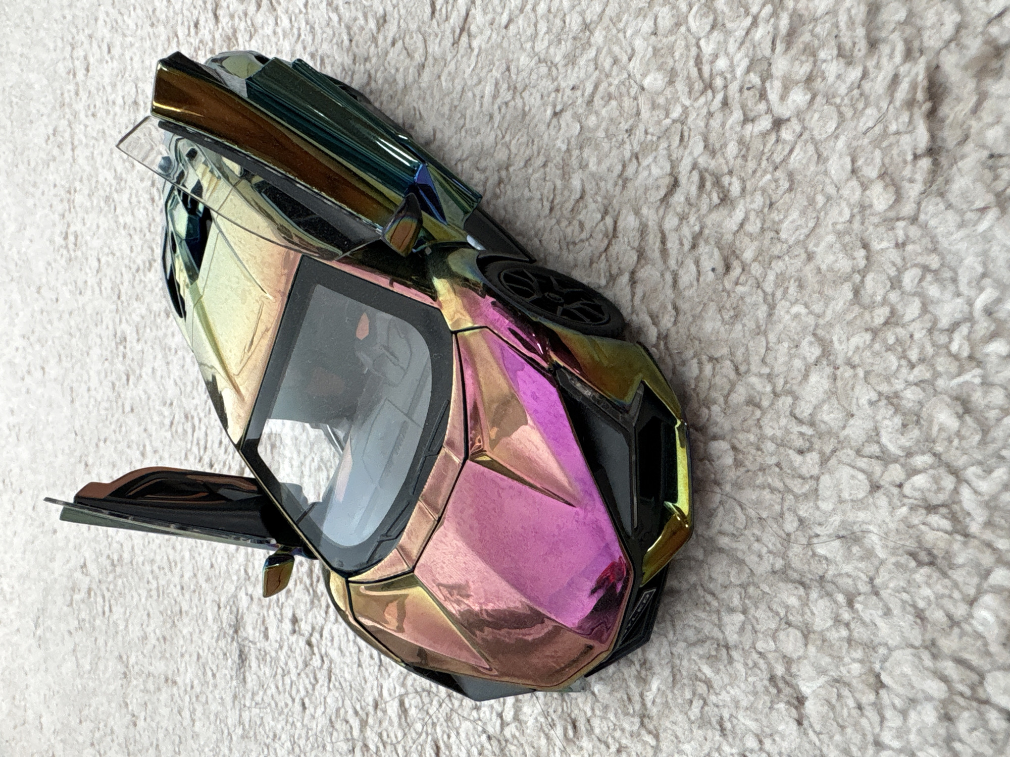 A detailed overhead shot captures an iridescent toy model sports car, featuring a striking chameleon paint job, resting on a textured, light-colored, shaggy carpet in Sofia, Bulgaria. The car's metallic finish displays a vibrant color shift, with the front hood appearing primarily pink and magenta, transitioning to golden-orange hues along the front fenders and then to iridescent green and blue along the sides and rear. Both of the car's distinctive scissor-style doors are open and angled upwards. The interior of the car is dark. The wheels, lower front bumper, and side skirts are black, and a small inscription "TYPER" is visible on the front. Small, dark strands, resembling hair, are visible on the carpet surface in several areas, including a prominent wavy strand in front of the car's nose. The overall scene is brightly lit, highlighting the car's reflective surface and the carpet's texture.