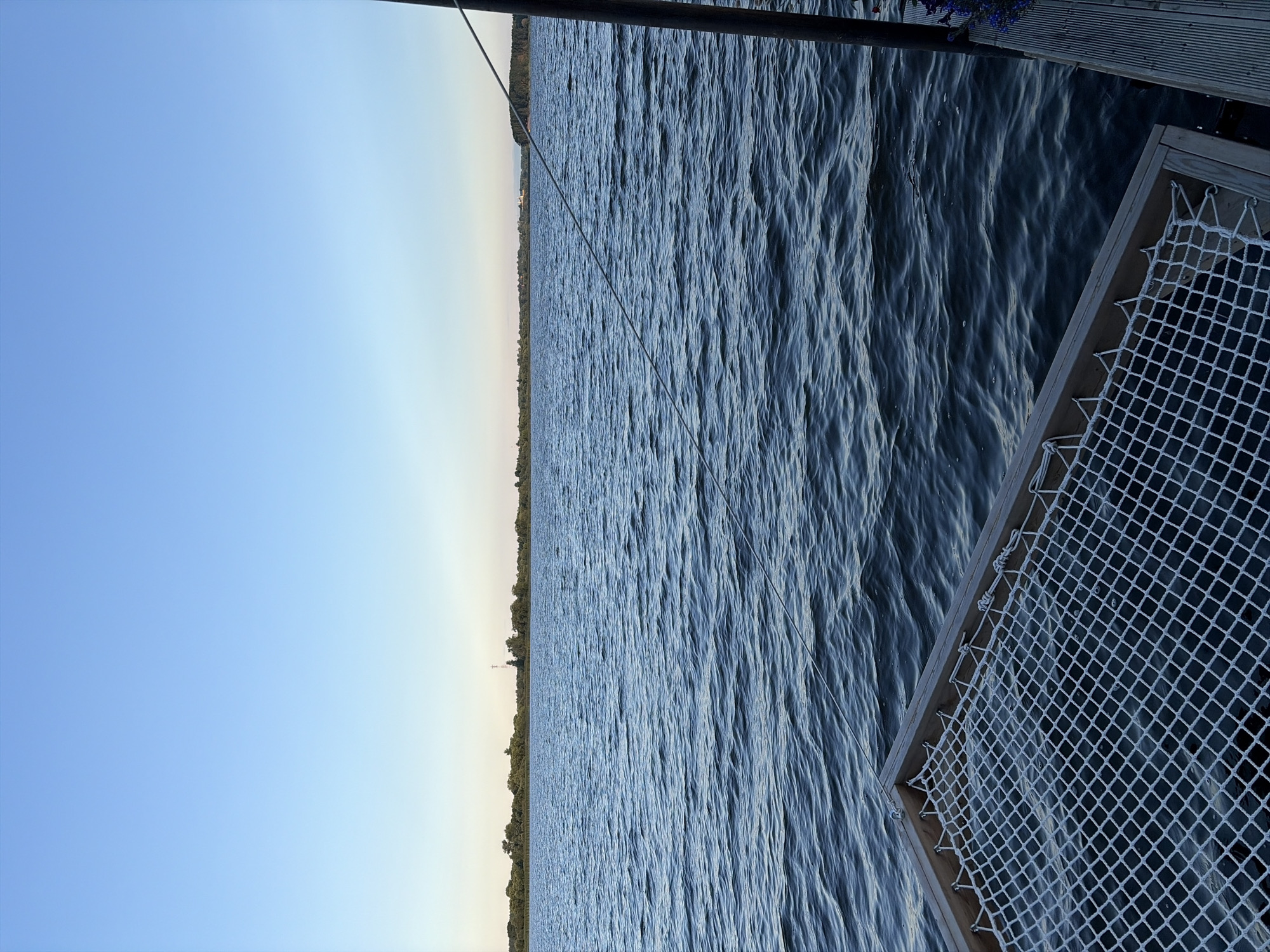 This is a vertical, eye-level, outdoor photograph of a body of water with a clear sky. In the foreground, a wooden frame with a white net, likely part of a hammock or a floating dock, is visible. The net appears to be suspended over the water. A thin rope extends diagonally from the upper right of the frame towards the lower left, connecting to unseen structures. The water is choppy with small waves and reflects the blue sky. In the mid-ground, a treeline with scattered structures, including what appears to be a radio tower, forms the horizon. The sky is a gradient of light blue, lighter towards the horizon. The location is identified as Vidogoshchi, Russia.