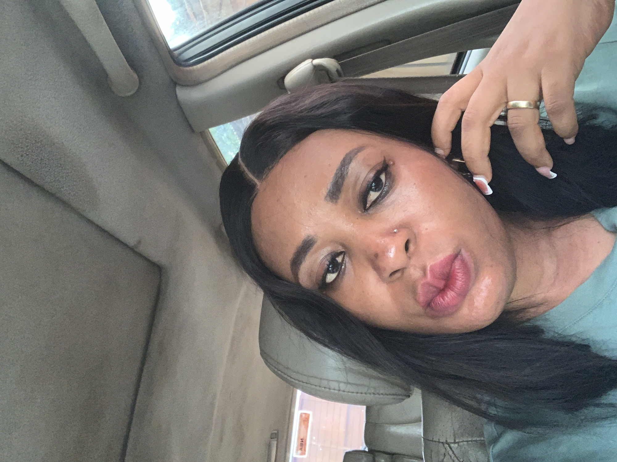 A woman is inside a car. The location is unknown and there is no information to suggest it is Sabon Tasha, Nigeria.
The woman is wearing a wig, a gold ring, and a light green shirt. She has a nose piercing and light colored nail polish. She is holding the wig near her ear. The interior of the car is gray.