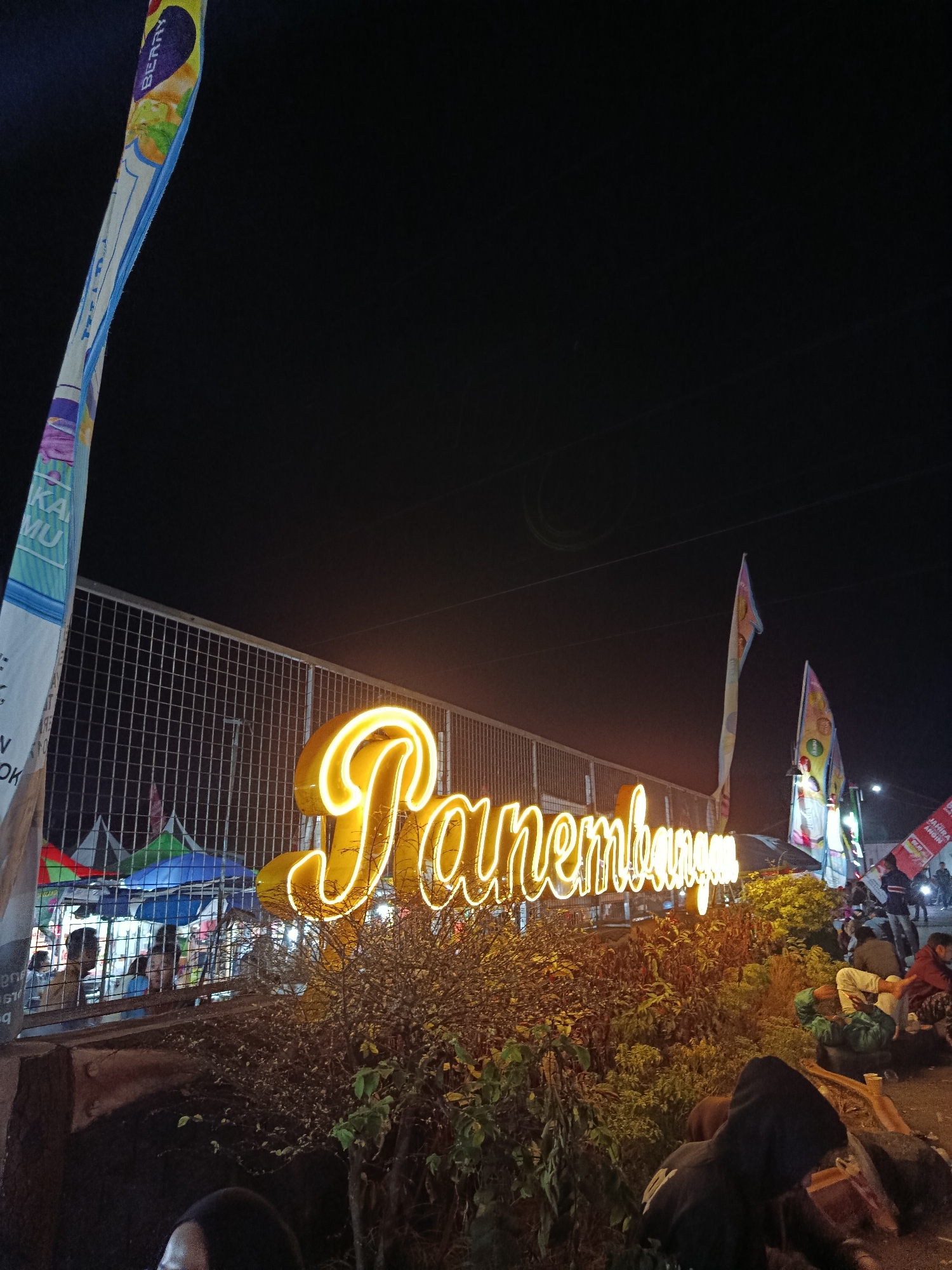 The image depicts a nighttime scene at an outdoor market or event in Panembangan, Indonesia. A large, glowing neon sign, likely spelling out "Panembangan" or a related phrase in cursive, dominates the center of the frame, emitting a warm yellow light. The sign is set against a backdrop of a wire fence and a dark night sky.
In the foreground, several people are visible, some seated on the ground, appearing to be resting or observing the surroundings. A person wearing a dark hoodie is in the lower right corner, facing away from the camera. To the left, a few people can be seen behind the fence, possibly interacting at stalls or booths.
Various flags with colorful patterns and text are planted around the area, adding to the festive atmosphere. Some tents or market stalls are visible behind the fence on the left side of the image. The ground appears to be a paved surface, and there is vegetation, including bushes and small trees, around the neon sign. The overall impression is of a lively evening gathering or celebration.