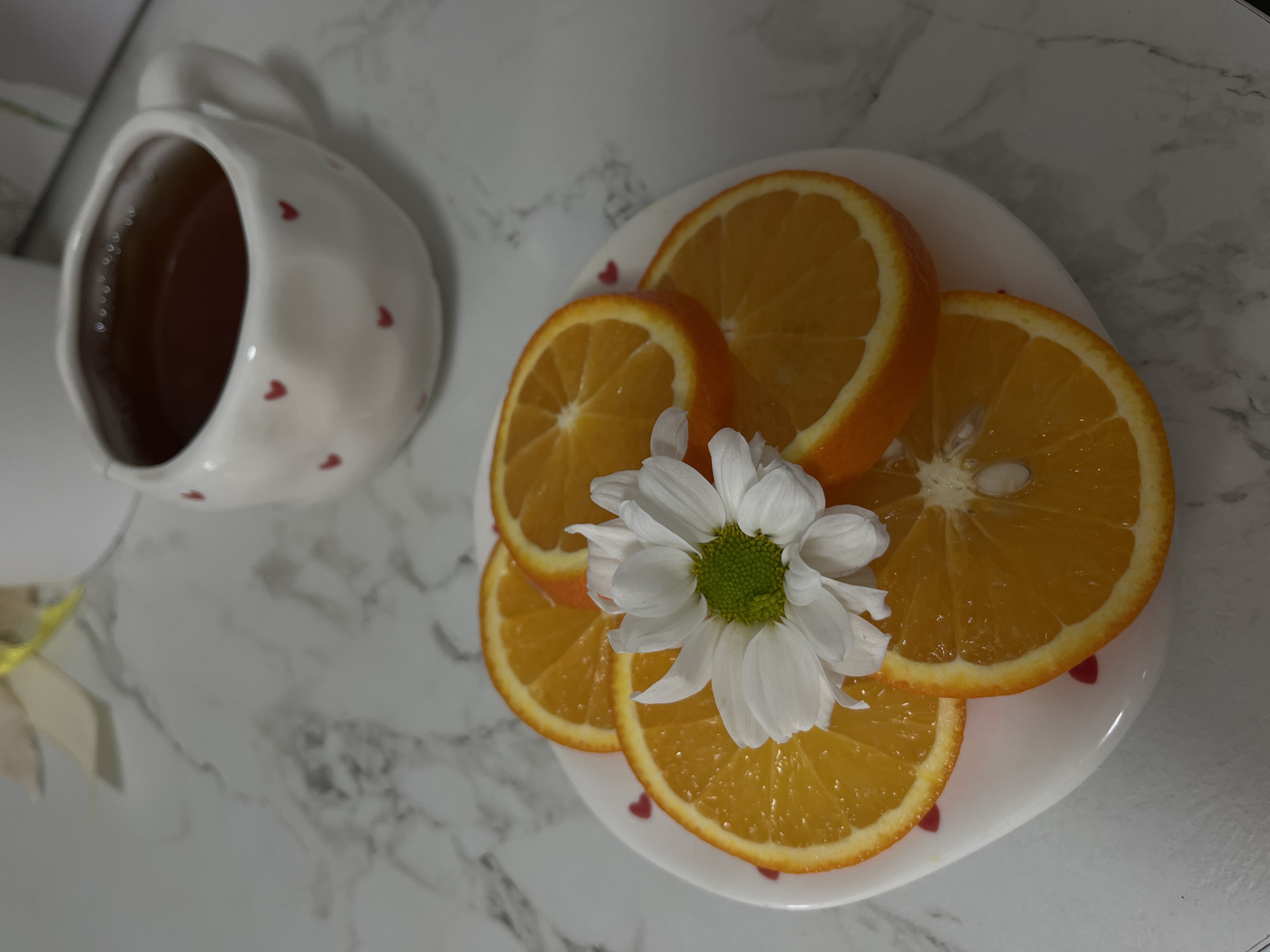 The image shows a white ceramic mug with small red heart decorations, filled with a dark liquid, positioned next to a white plate on which are arranged several slices of orange with a white daisy flower on top. The plate also has small red heart decorations. The surface beneath these objects appears to be a marble-patterned countertop. There is no indication that the image was taken in Chaikovsky, Russia. There are no people visible in the picture.