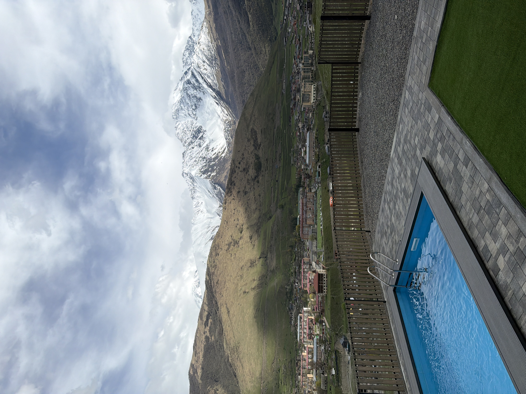 A high-angle view depicts a bright blue swimming pool in the foreground, positioned near a paved area with gray rectangular stones, artificial green turf, and dark gravel. A wooden fence separates the pool area from a densely built-up town, nestled in a valley surrounded by large mountains. The lower parts of the mountains are covered with brown and green vegetation, while the higher elevations are capped with snow. The sky is partially covered with clouds. The scene is Verkhny Fiagdon, Russia.