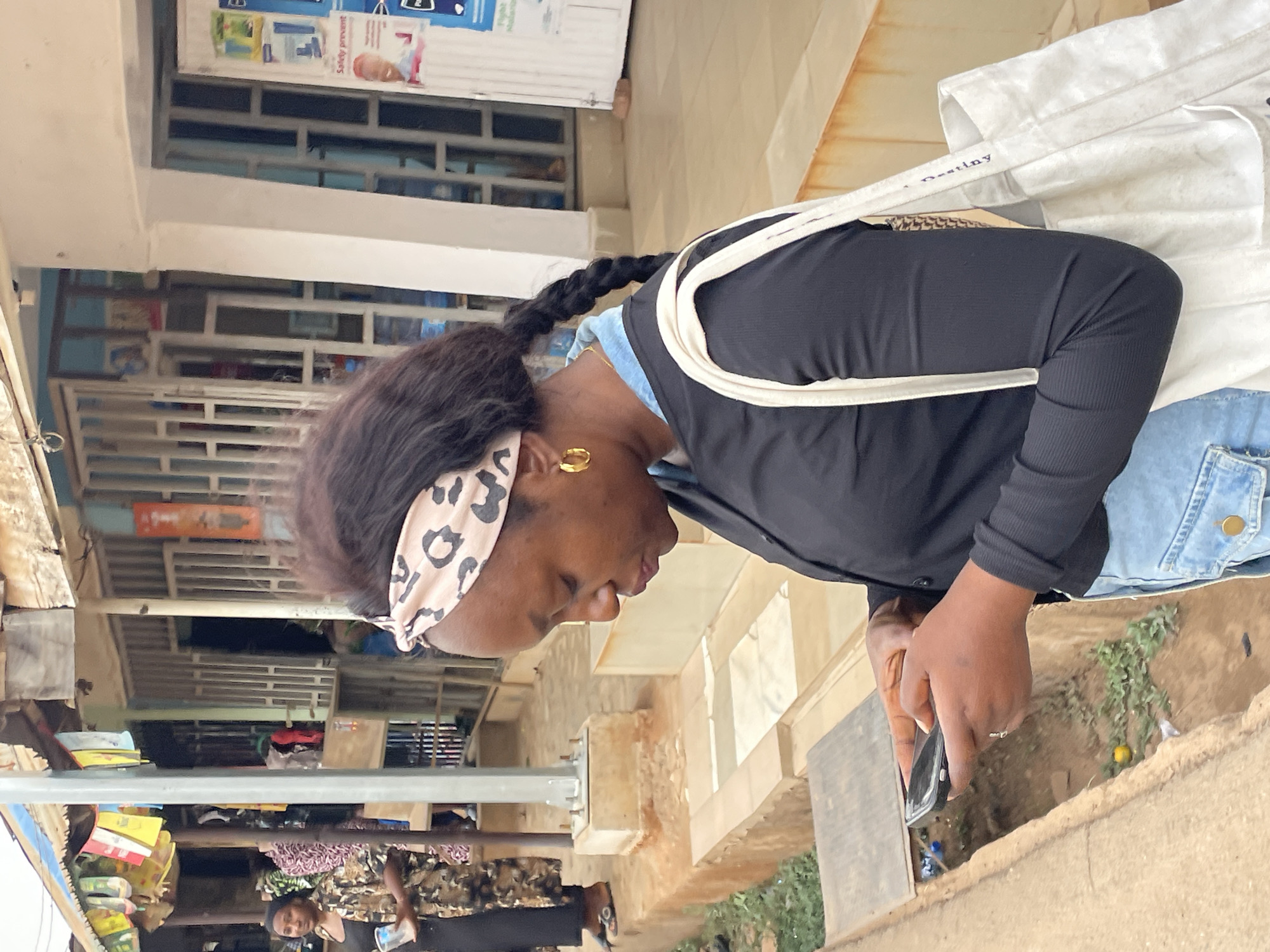 A woman wearing a black long-sleeved shirt, a leopard print headband, and denim pants is looking at a phone in her hands. She is in an urban environment, identifiable by the architecture. In the background are market stalls, and signs are visible on some of the shops. Additional people can be seen in the stalls. The location can be identified as Jos, Nigeria based on architectural style and the market atmosphere.