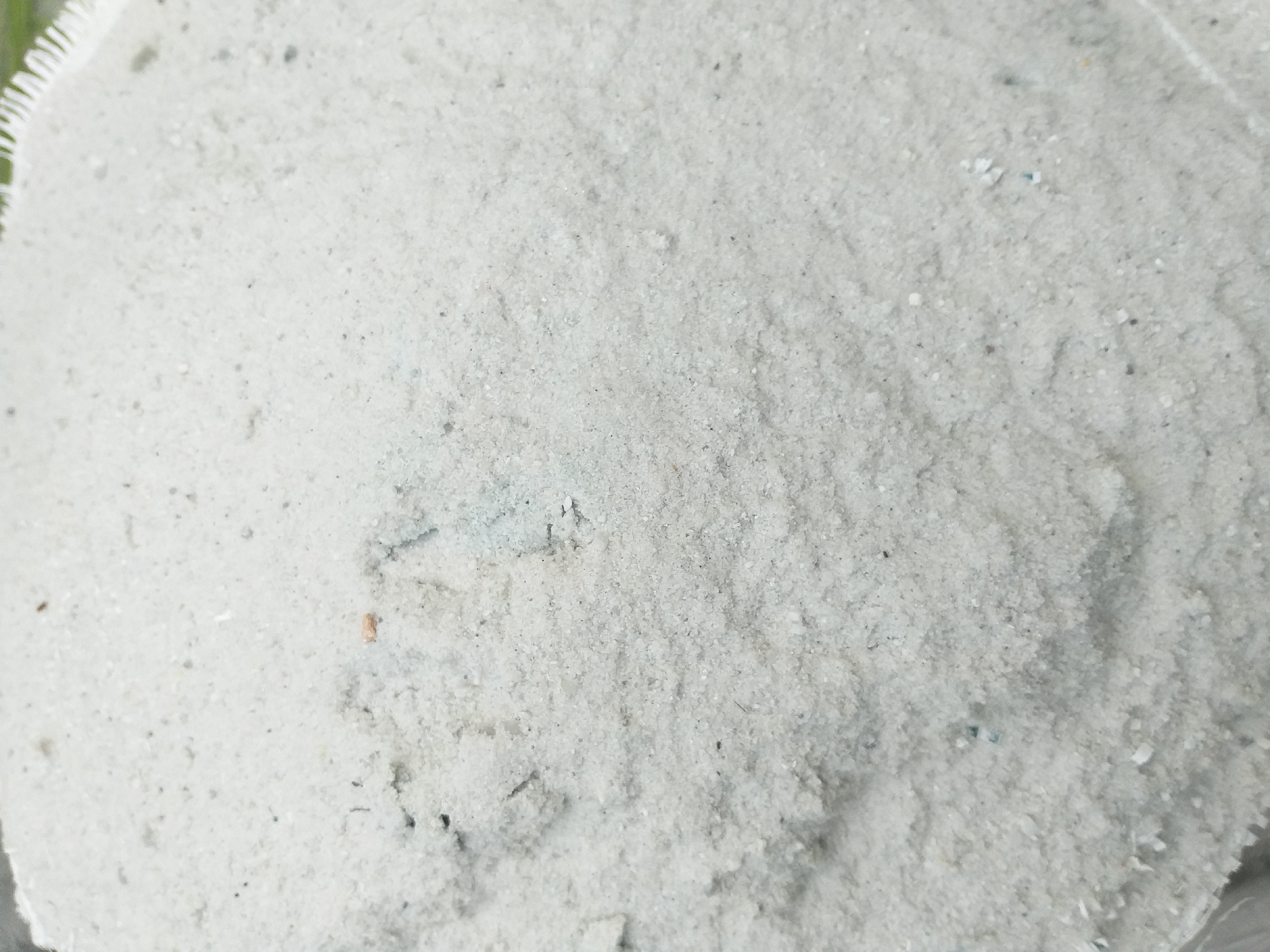 The image shows a close-up, overhead view of a pile of light-colored sand, likely construction sand, with some minor debris mixed in. The sand appears slightly damp and is piled in a way that creates undulating textures and shadows. In the upper right corner, a section of a woven white material with a fringed edge is visible, possibly the top of a bag or tarp. The overall impression is of a material storage area. The location in Air Jamban, Indonesia is not discernible from the image itself, as there are no specific landmarks or contextual clues.
