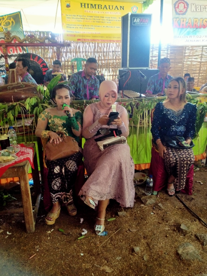 Three women in formal attire are seated in front of a bamboo backdrop, while several men play instruments in the background. The woman on the left holds a microphone, the middle one uses a mobile phone, and the woman on the right holds money. A speaker is behind the musicians. A banner reads "HIMBAUAN PERATURAN BERSAMA". Another banner reads "KHARIS BALON RT.02/ JENAWI, KA". The ground is littered with debris.