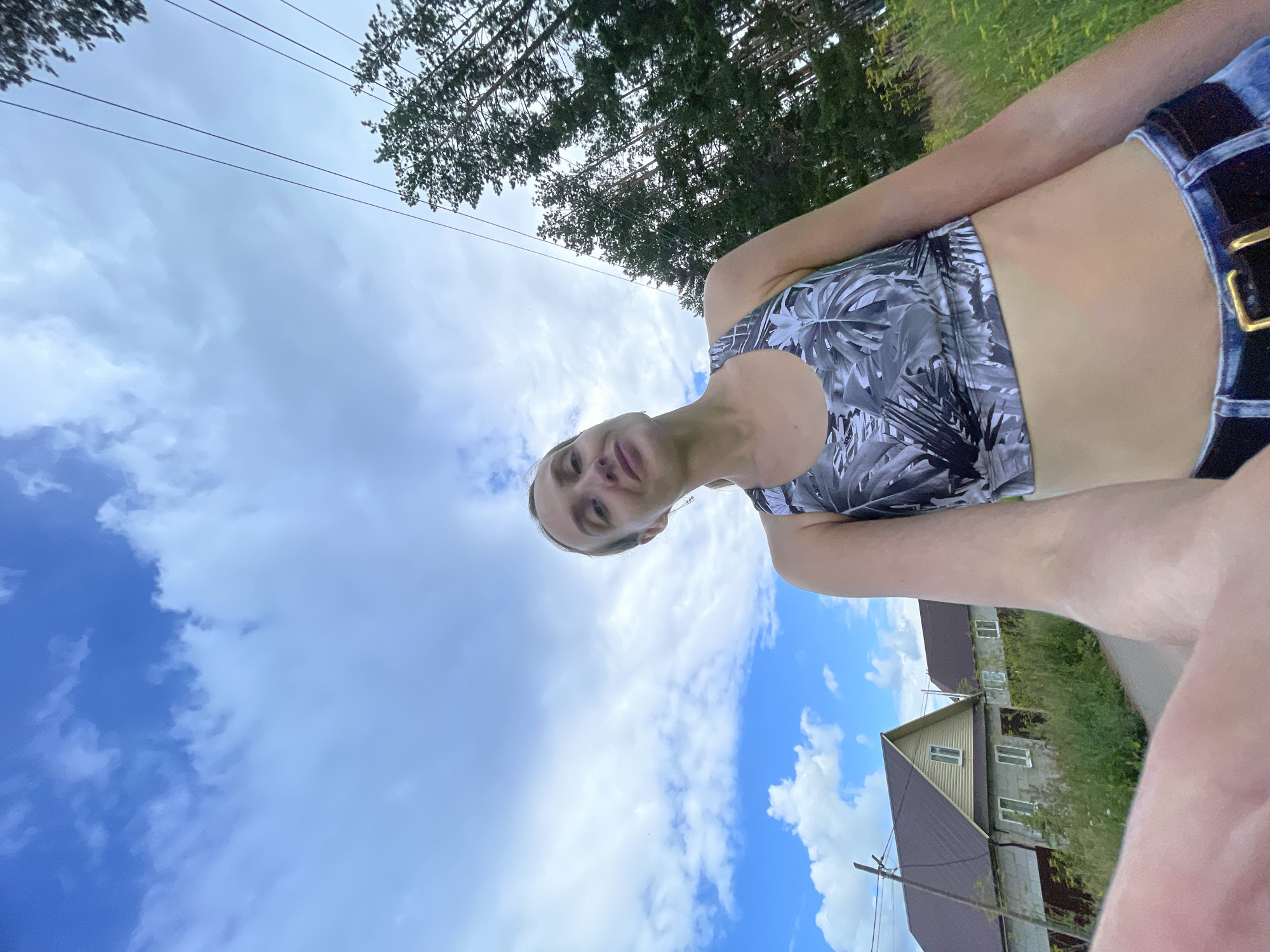 A woman is taking a selfie outdoors, holding the camera with her left arm, which is extended towards the viewer. She is wearing a black and white patterned sports bra and blue denim jeans with a gold-buckled belt. Her right shoulder and upper arm are visible on the right side of the frame. The background features a blue sky with scattered white clouds, tall green trees on the right, and two houses with brown roofs and light-colored walls on the left. A dirt road or path is visible in the foreground, and power lines run diagonally across the sky. The scene appears to be in a rural area, possibly the village of Podshivalovo, Russia, given the architectural style of the houses and the presence of a dirt road and power lines.