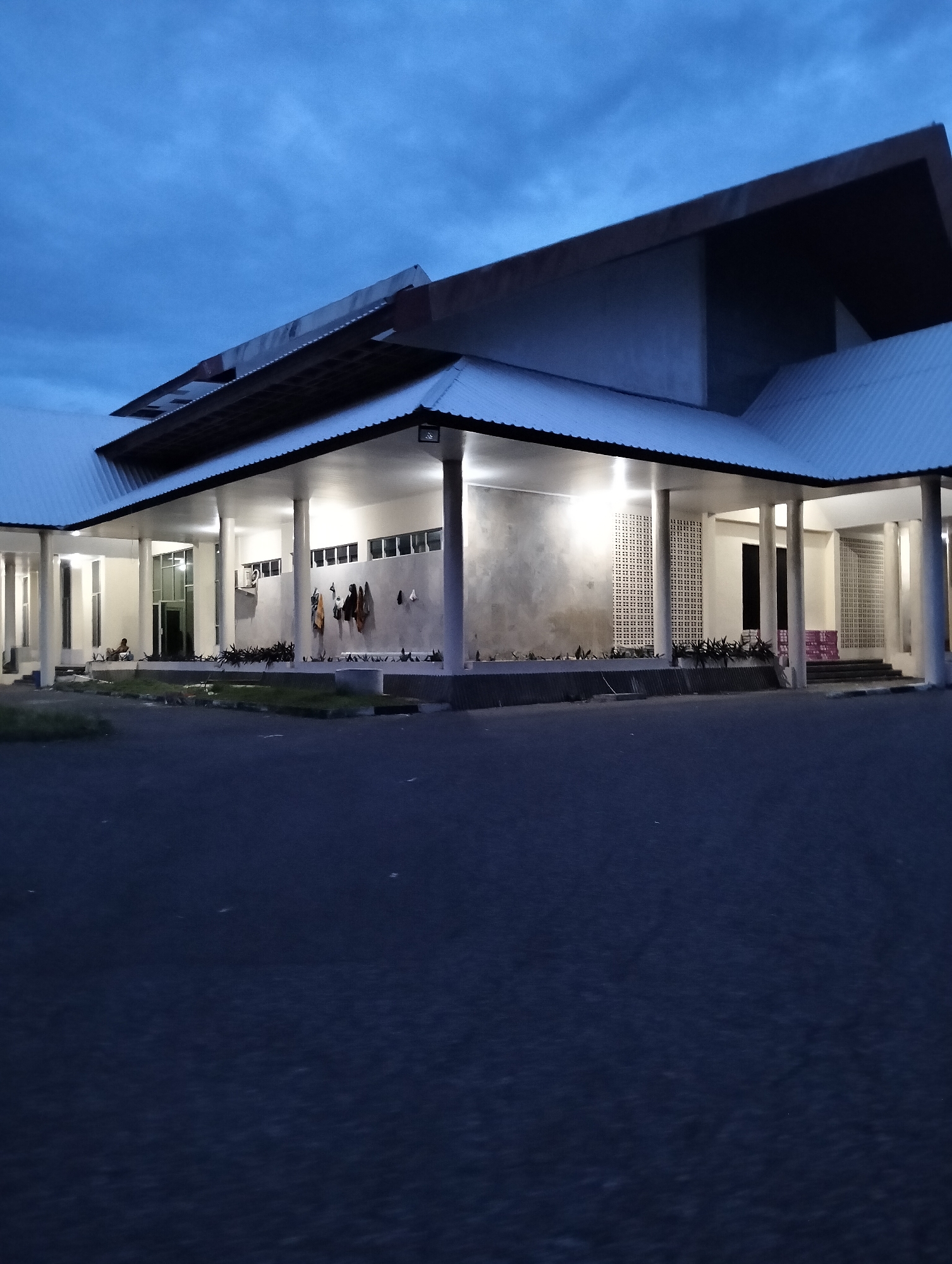 The image shows a modern building with a wide, covered walkway supported by columns. The building appears to be a public facility, possibly a community center or a place of worship, given the presence of what looks like prayer mats laid out on the ground. Several pieces of clothing are hanging on a wall in the covered area. A single person is sitting outside the building. The sky is dark blue, indicating it is either dusk or dawn. The scene is set in Palohgadeng, Indonesia.