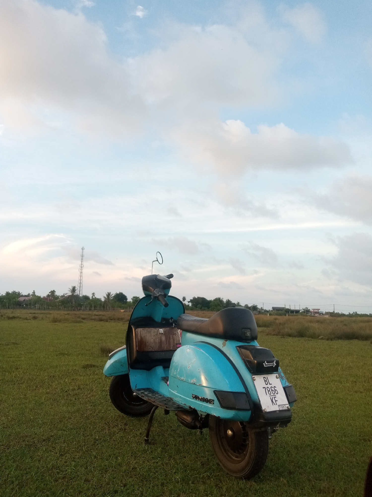 A light blue Vespa scooter is parked on a grassy field in Bie, Indonesia, under a cloudy sky. The scooter is facing away from the viewer, with its rear visible. A license plate reading "BL 7866 KF" is attached to the back. In the distance, a cell tower and some buildings are visible amidst a line of trees. The field is unevenly mowed and shows signs of dry patches. The overall lighting suggests it is late afternoon or early evening.