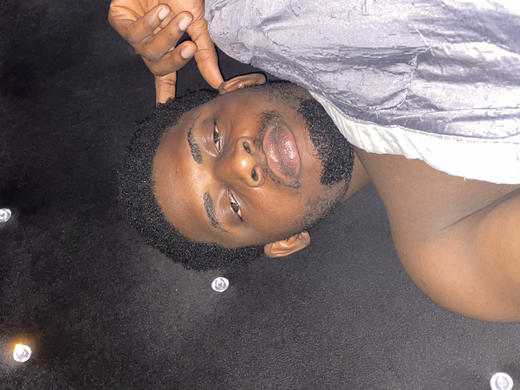 A dark-skinned man is pictured lying on his back, looking directly at the viewer. He has short, curly black hair and a well-trimmed beard and mustache. His eyes are open, and his mouth is slightly ajar, revealing moist, full lips. His left hand, with fingers slightly curled, rests near his head, with one finger touching his hair above his ear. He is wearing a light-colored, possibly grey or silver, textured garment that appears to be a shirt or jacket, with a visible white trim along its edge. The man is positioned on a dark, textured surface, likely a floor or carpet, which appears black. Scattered on this dark surface are several small, shiny, reflective objects, resembling decorative studs or embedded jewels. The lighting is soft, highlighting his features. His expression is neutral and calm. There is no visible text in the image. The scene suggests an indoor setting, and the activity is a posed portrait while lying down.