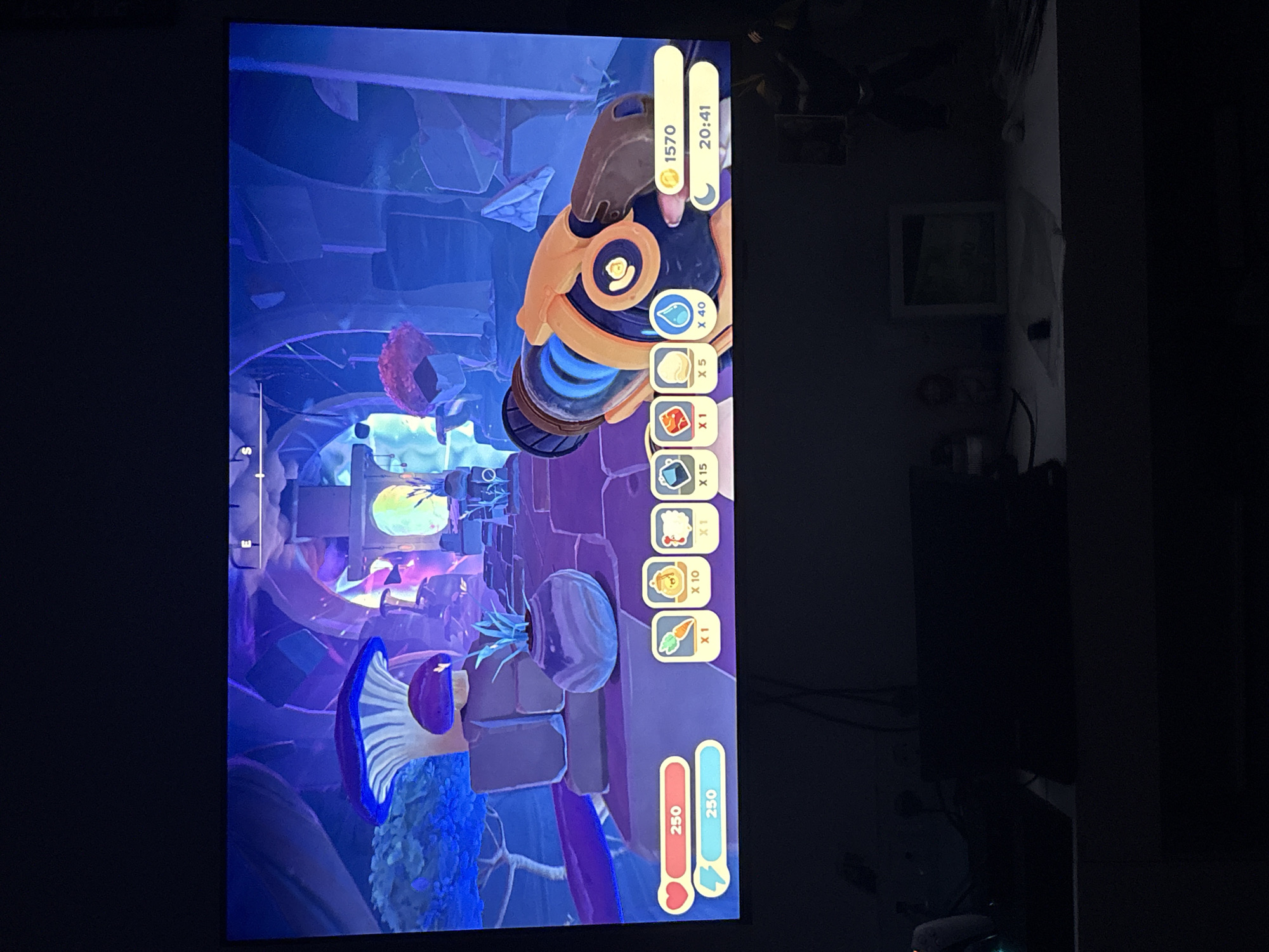 The image depicts a vertically oriented display screen, illuminated and showing a video game interface. The game features a vibrant, whimsical environment with purple, blue, and yellow hues. Dominating the right side of the screen is a graphical user interface showing in-game currency (1570), time (20:41), resource quantities (water x40, wheat x5, meat x1, sap x15, wool x1, honey x10, carrot x1), and character stats (health 250/250, energy 250/250).
The physical setting is a dark interior room. A dark television is visible and a piece of framed art on the wall, but these background elements are largely obscured by shadow.
There is no visible evidence in the image to suggest that this is located in the city of Plovdiv, Bulgaria.
