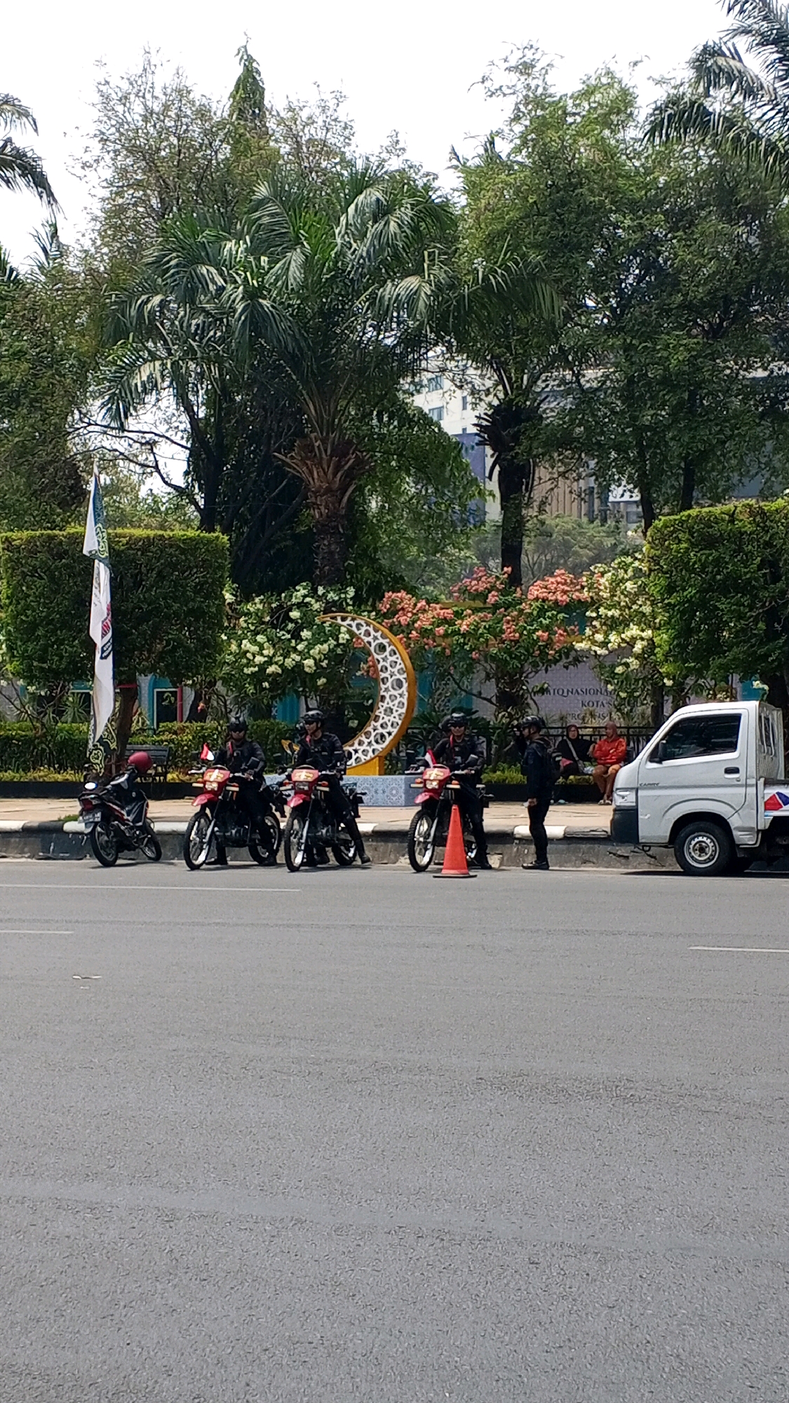 The image captures a street scene in Semarang, Indonesia, characterized by a lush green environment and a clear sky. The foreground is dominated by a gray asphalt road, marked with white lines. To the left of the road, a row of motorcycles is parked, with four riders in dark uniforms, some wearing helmets, seated on their vehicles. Behind the motorcycles, a decorative crescent moon sculpture, adorned with white and pink flowers, stands prominently against a backdrop of dense green foliage and trees. Further back, a white pickup truck is visible, parked on the right side of the scene. Two individuals, a woman in an orange jacket and a person in dark clothing, are seated on a bench behind a low hedge, observing the surroundings. The overall ambiance suggests an outdoor public space, possibly a park or a ceremonial area, in a tropical urban setting.