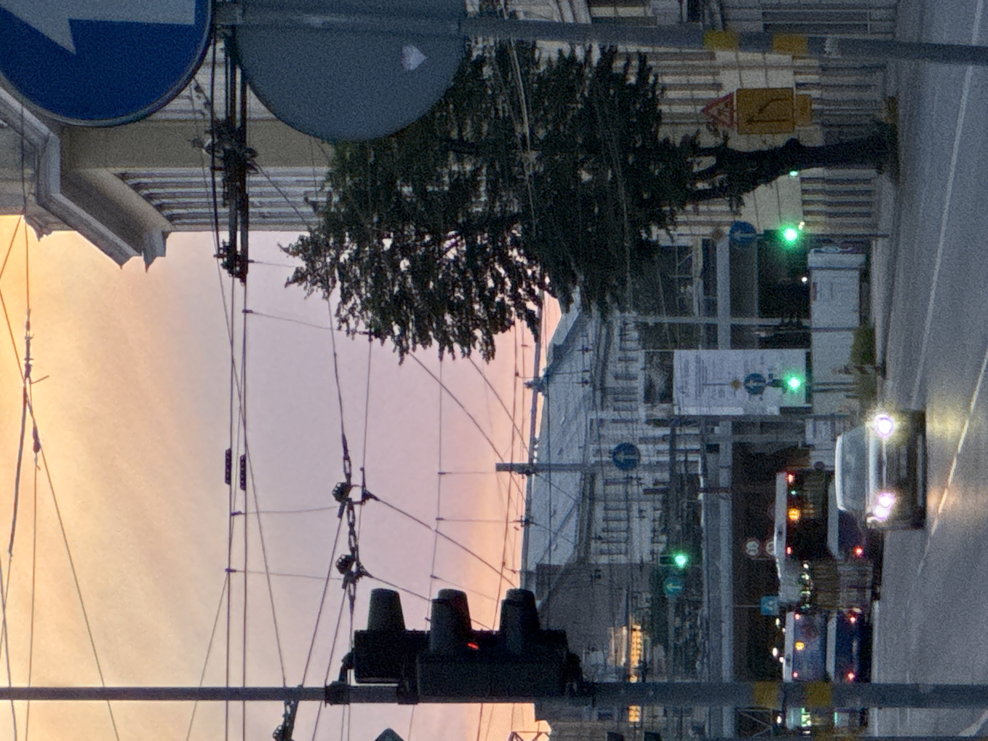 The image depicts a street scene in Riga, Latvia during twilight, with a soft, peachy-pink sky. Overhead, a complex network of electrical wires for trolleybuses crisscrosses the frame. In the foreground, a traffic light on a pole shows a red signal. Further down the street, two buses are visible, one in the left lane and another slightly ahead in the right lane, both with illuminated headlights and taillights. A car with its headlights on is in the right lane, approaching the viewer. Several buildings line the street, some with visible architectural details and windows. A tree stands prominently on the right side of the road, and traffic signs are also present. The scene appears to be an urban environment with ongoing traffic.