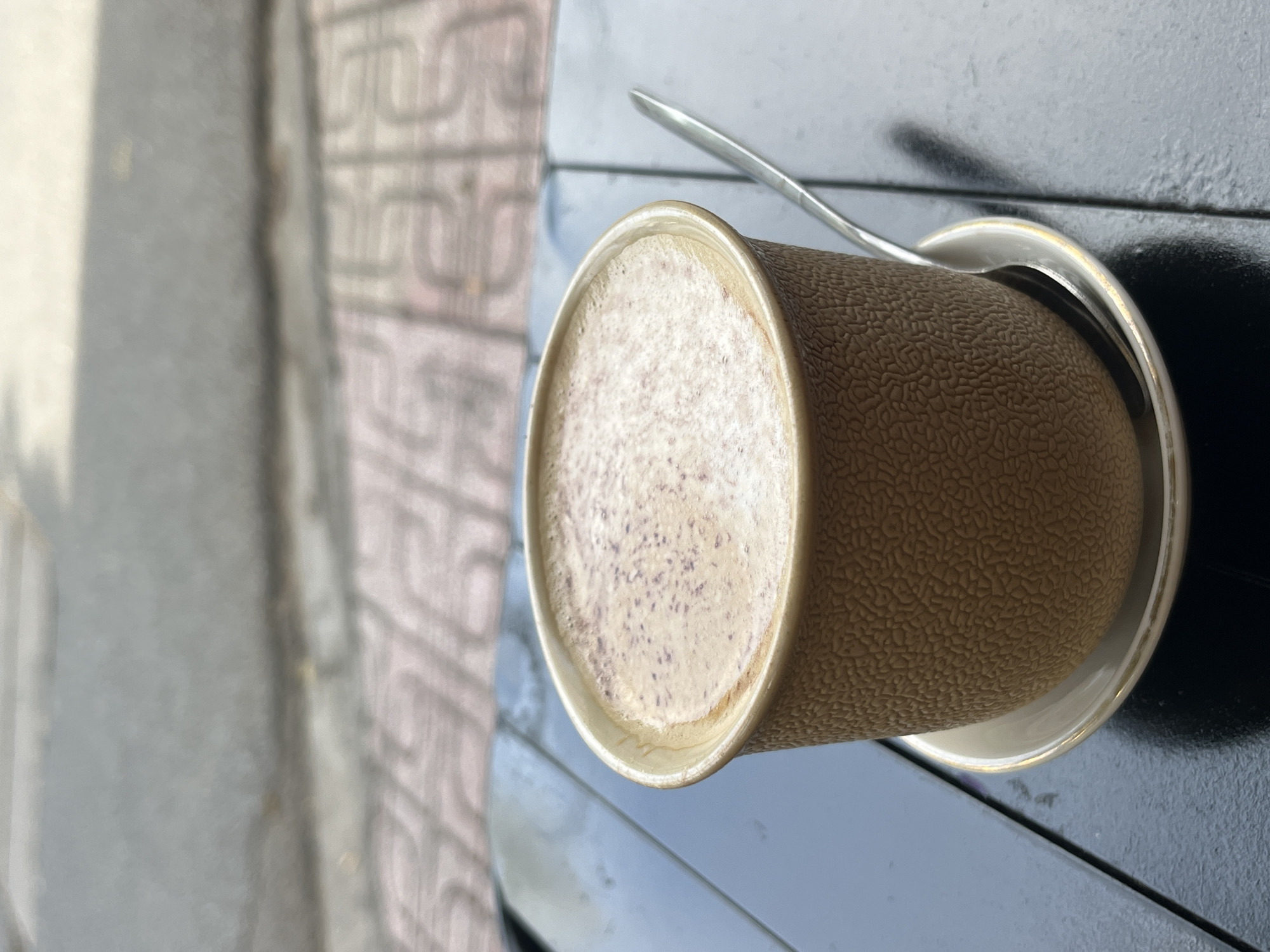 A close-up, eye-level shot shows a textured, tan-colored cup of coffee on a saucer with a silver spoon resting on the saucer. The cup is filled with coffee topped with foam, dusted with cinnamon. The coffee cup sits on a dark, reflective surface, likely a table, with a blurred background of a city sidewalk in Nha Trang, Vietnam. The sidewalk is made of patterned light-colored tiles, with a grey concrete border. The overall lighting suggests an overcast day. There are no people visible in the image.