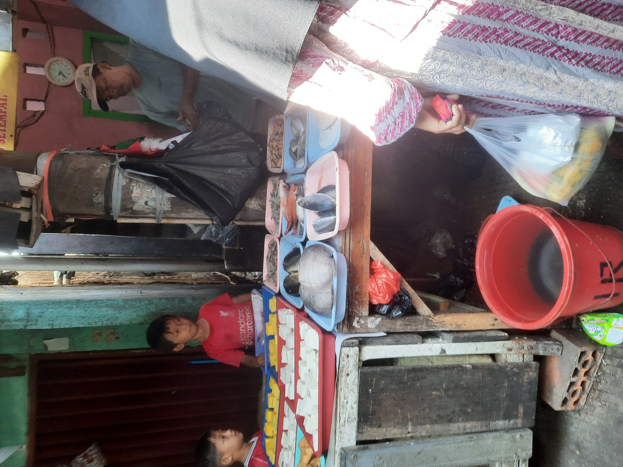 In North Jakarta, Indonesia, an outdoor market scene unfolds. Two young boys, one in a red t-shirt with "Standard Chartered" branding and the other partially visible in a red shirt, stand near a display of food items. Their gaze is directed towards the food. Adjacent to them, a man wearing a green shirt and a tan cap is visible, appearing to be engaged in preparing or handling goods.
In the foreground, a woman, partially obscured by her patterned clothing and hijab, holds a plastic bag filled with produce. A large red bucket, filled with dark liquid, sits on the ground near the market stall. The stall itself is constructed from weathered wood and features various trays filled with food, including tofu and several types of fish. Behind the stall, a black plastic bag is suspended. The environment suggests a local, informal market setting, likely with limited infrastructure.