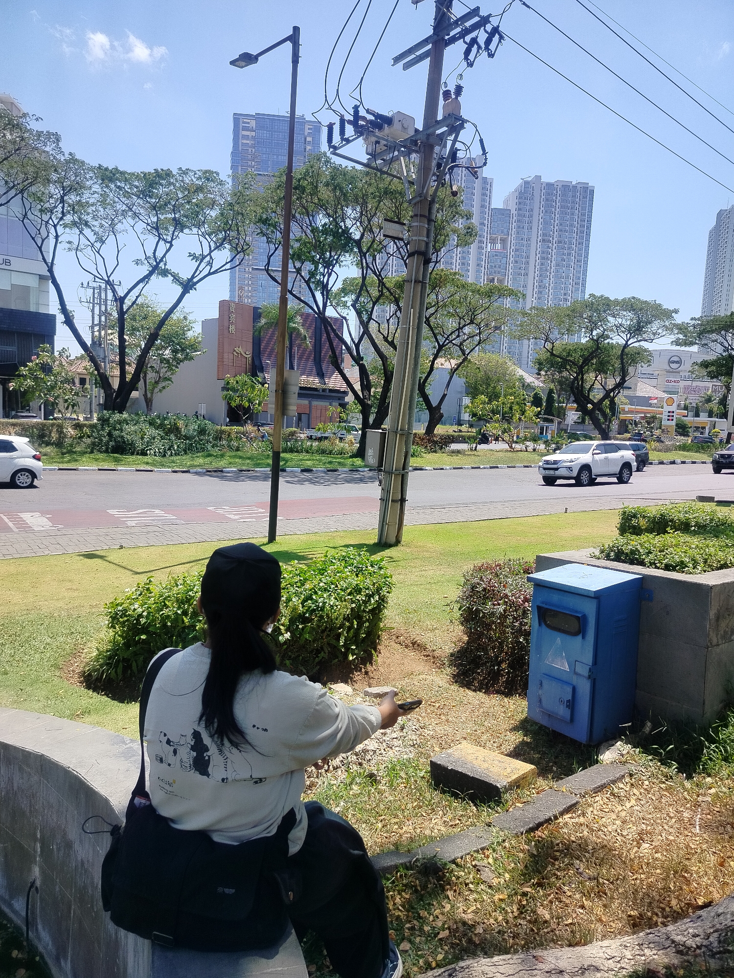 This image captures a sunny day in Surabaya, Indonesia. In the foreground, a person wearing a black cap and a light-colored sweatshirt with a graphic print sits on a concrete ledge, facing away from the camera. A black messenger bag is slung over their shoulder. The person appears to be holding a smartphone in their right hand, possibly taking a photo or using it.
The background reveals a vibrant urban scene. A paved road with traffic lanes separates the foreground from a manicured green median strip. Several cars are visible on the road, including a white SUV and a white sedan. Beyond the road, a row of trees with lush green foliage lines the street. In the distance, modern high-rise buildings stand tall against a clear blue sky. One of these buildings displays signage with the text "AZ-KO" and "SHELL," suggesting a commercial area with gas stations and retail outlets. A blue utility box is situated on the median strip near the person. The overall environment is characteristic of a well-developed urban area in Surabaya, likely a business or commercial district given the presence of high-rise buildings and branded establishments.